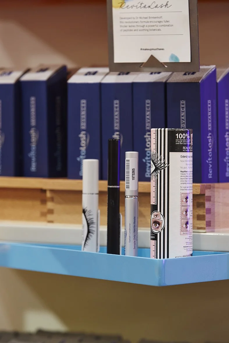 Assorted eye care products including mascara and eye drops displayed in a blue container on a store shelf, with a background of blue boxes and a framed informational sign.