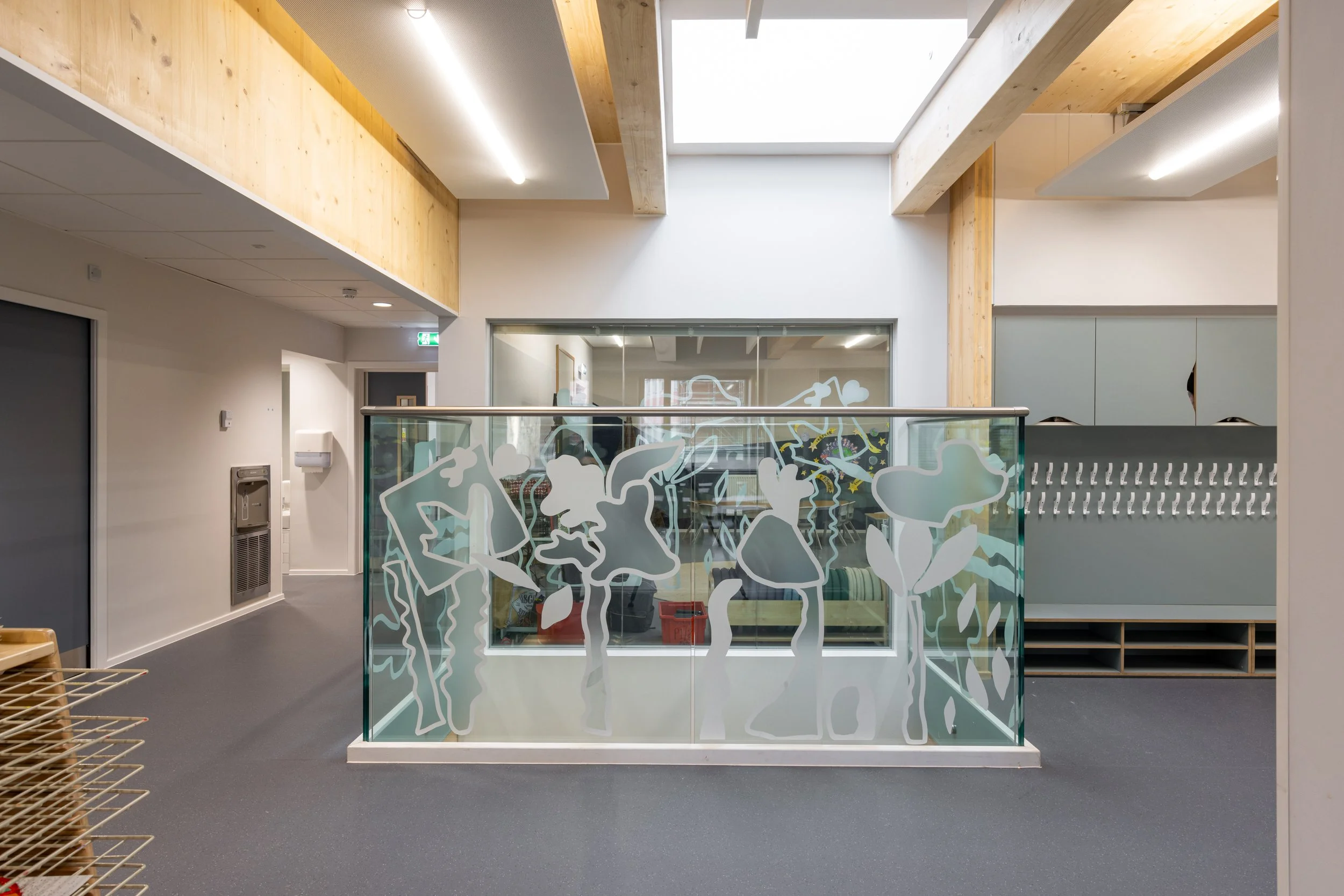 Interior view of a modern building with a glass railing decorated with abstract floral designs, a coat rack on the right, and a room with a large window and furniture in the background.
