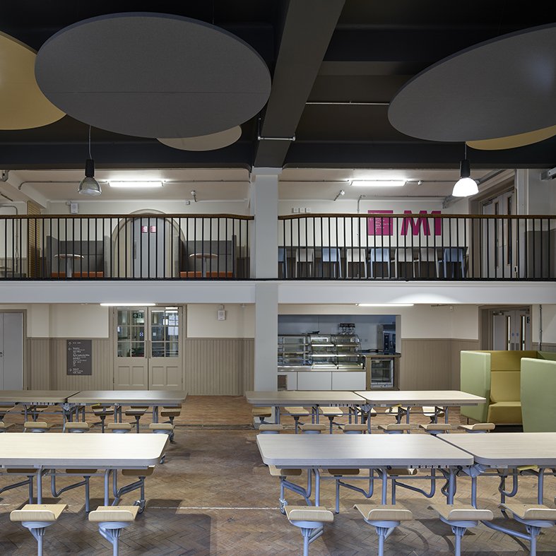 Empty cafeteria with folding tables and attached stools, upper balcony with seating areas and a pink 'M' sign on the wall.