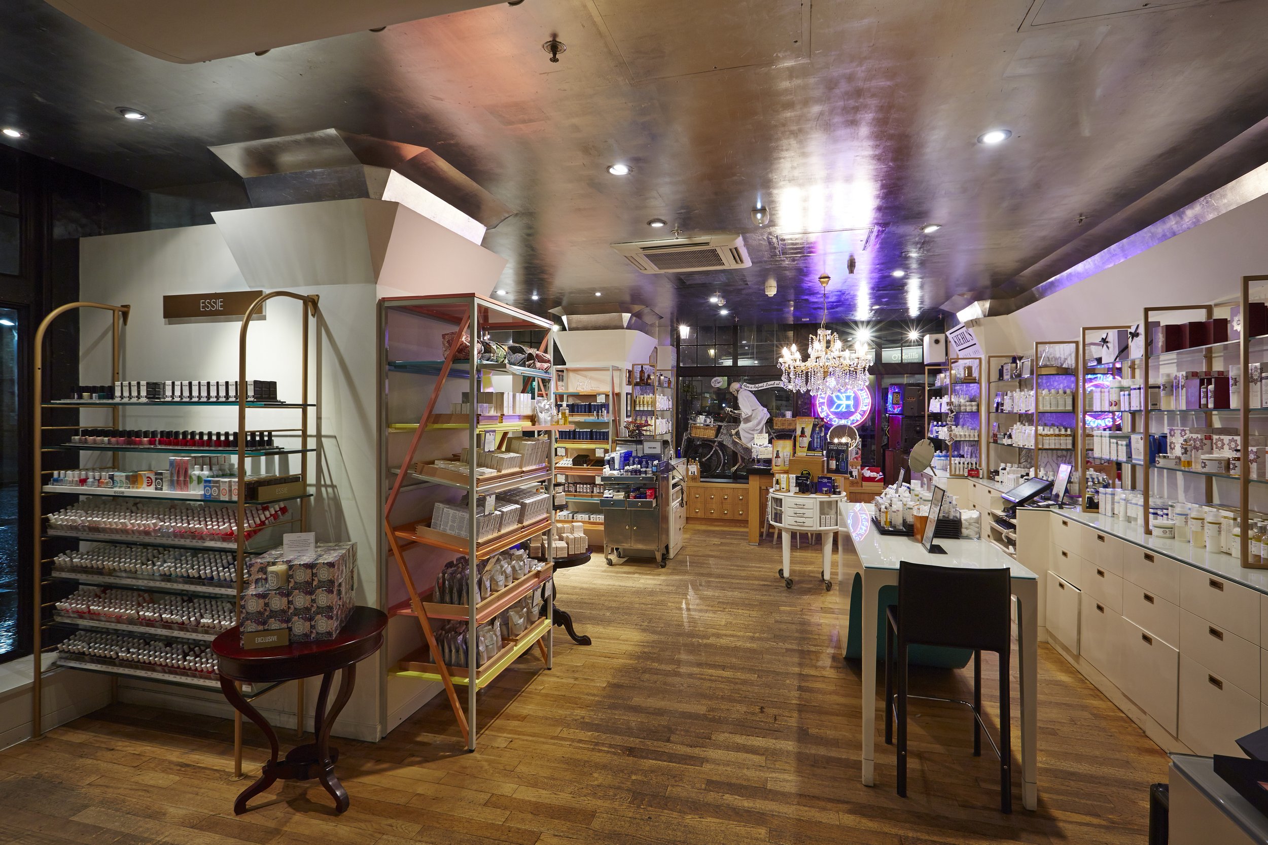 Interior of a boutique store with shelves stocked with cosmetic and skincare products, wooden flooring, and decorative lighting including a chandelier and neon signs in the background.