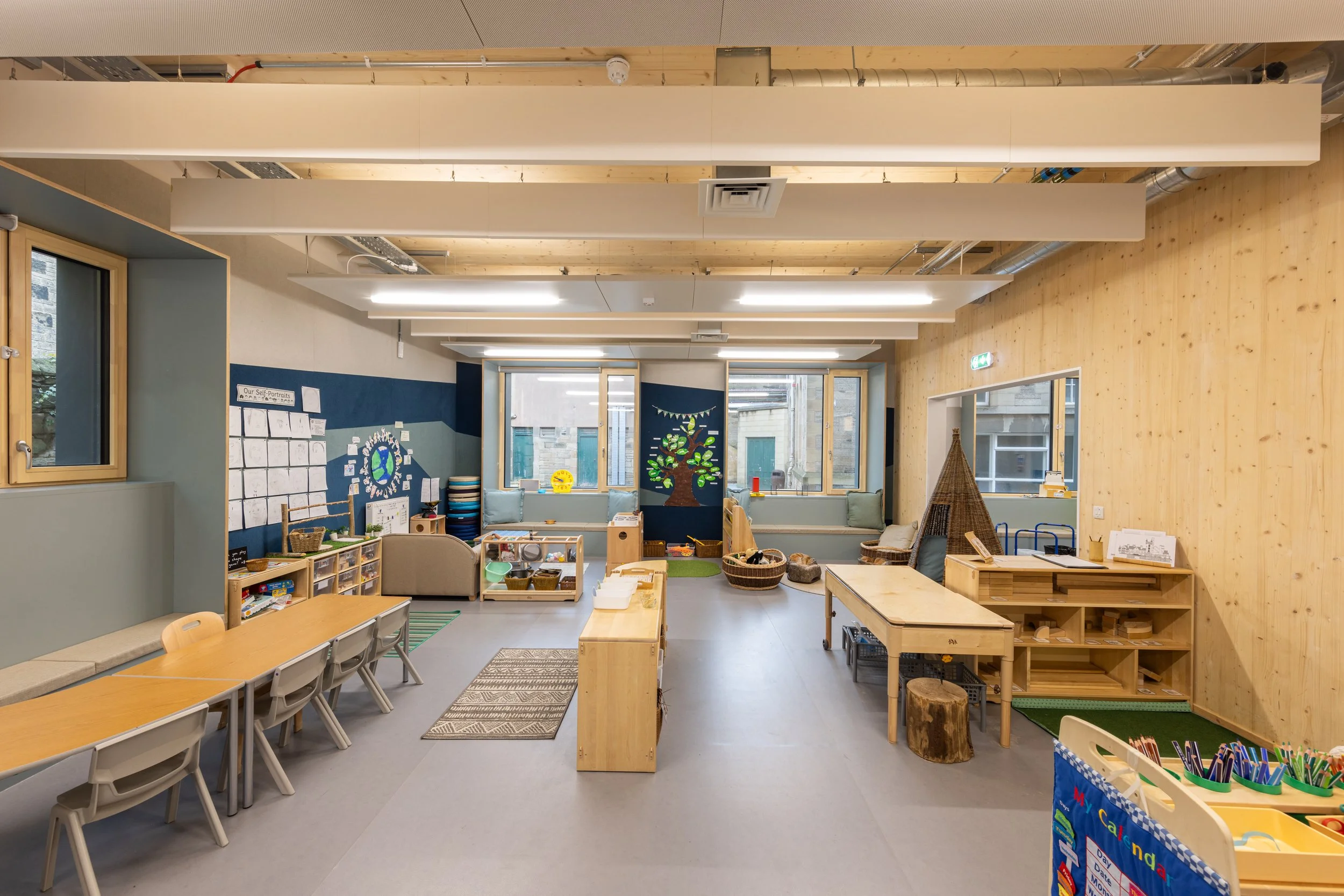 An empty classroom with wooden furniture, tables, chairs, and shelves, decorated with children's artwork and educational materials; natural light from windows illuminates the space.
