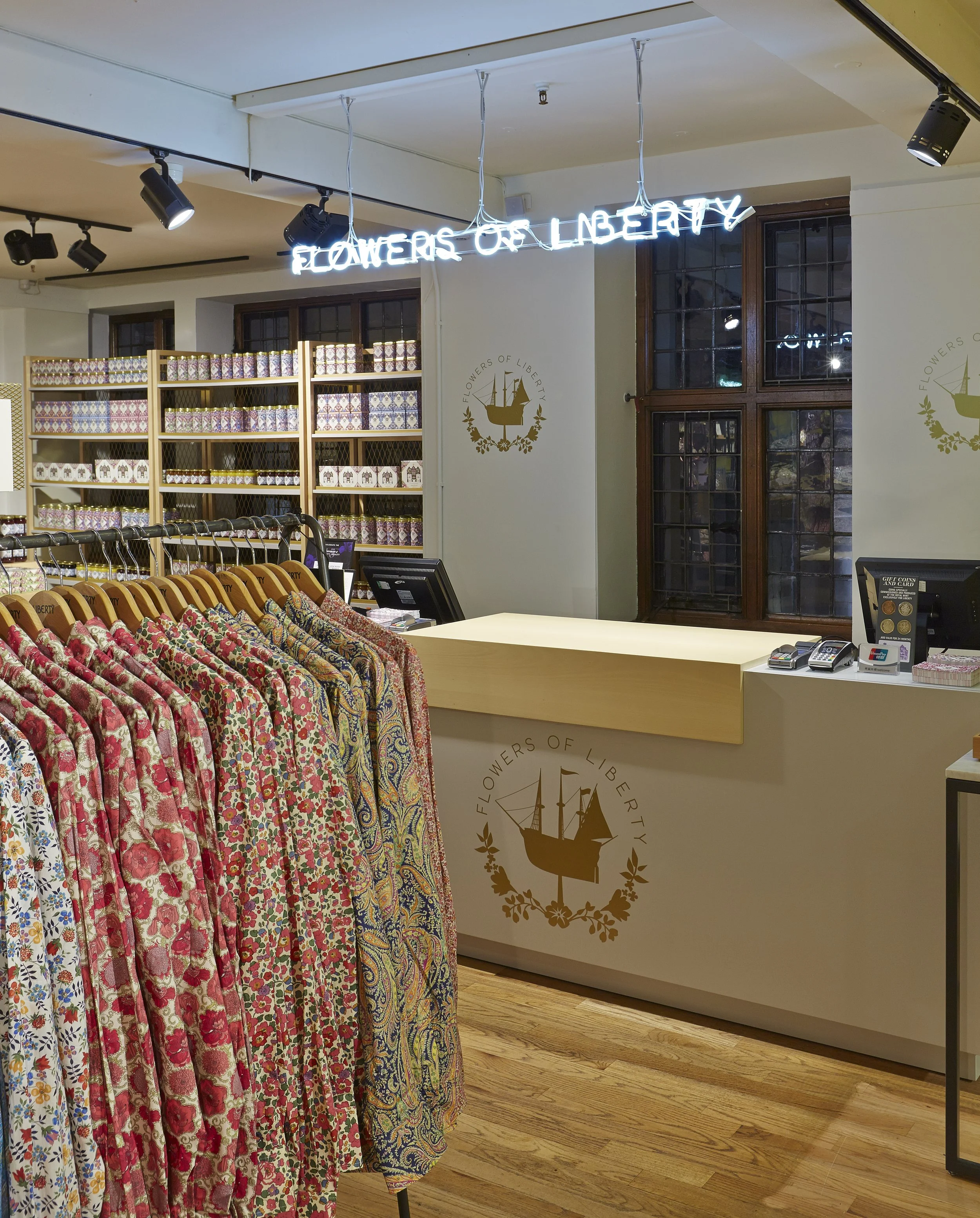 Interior of a store called Flowers of Liberty, featuring a clothing rack with patterned dresses, a wooden checkout counter with a logo, and shelves with floral-patterned boxes, illuminated by track lighting and a neon sign.