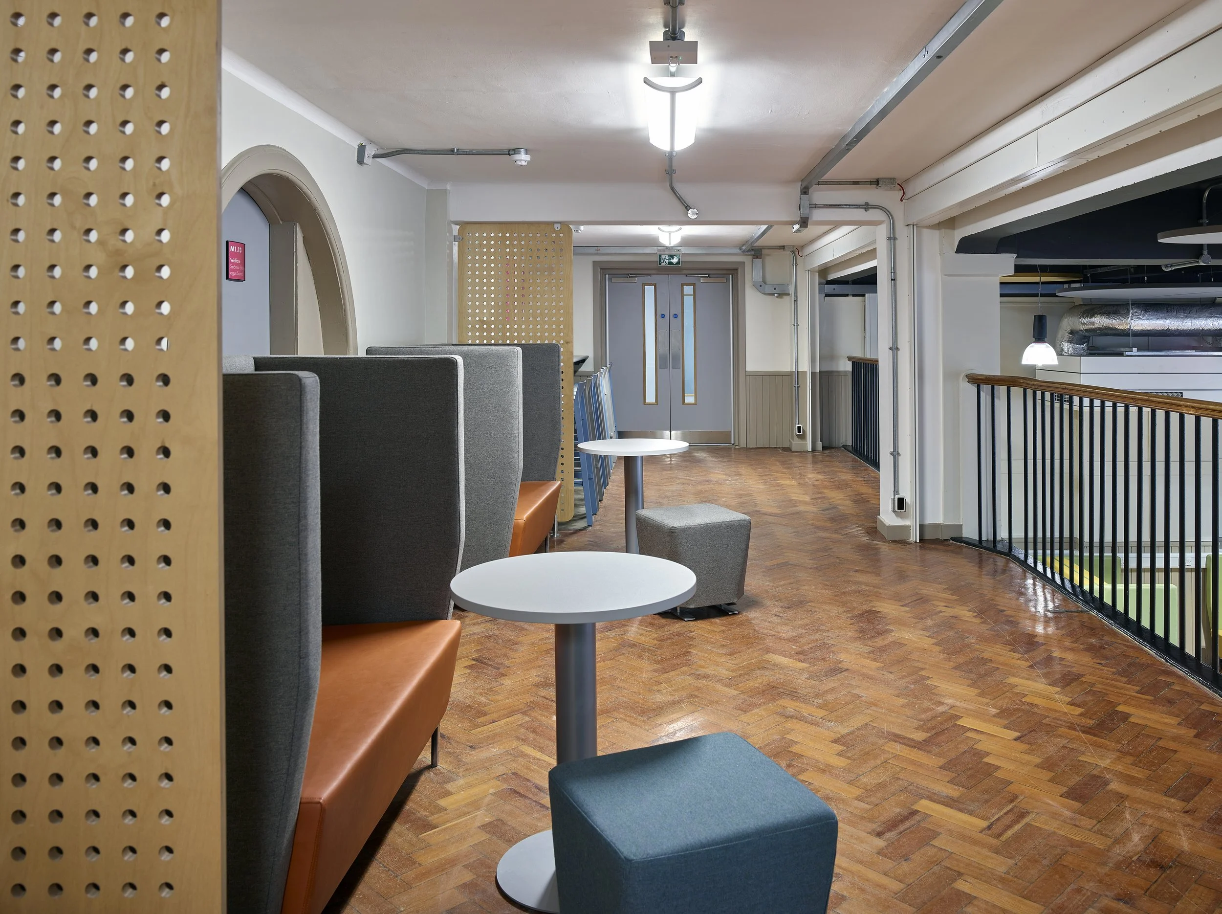 An empty modern lounge area with high back grey chairs, small round white tables, a blue ottoman, and wooden flooring. There's a railing to the right, some blue chairs stacked against the wall, and a doorway at the end of the room. Ceiling lights illuminate the space.