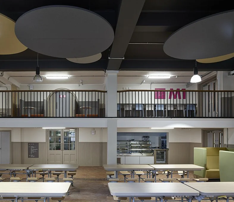 Interior of a modern cafeteria or dining area with high ceilings, round ceiling acoustic panels, a balcony with black railing, tables and chairs below, and a kitchen area with food serving equipment in the background.