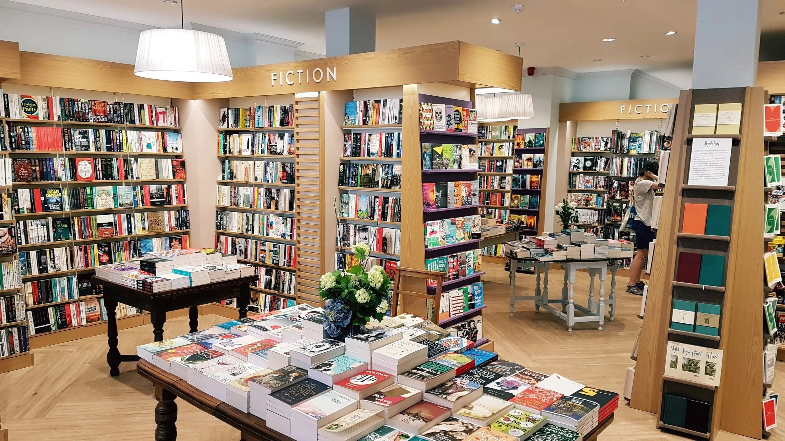 A bookstore with shelves of fiction books, display tables with books and a flower arrangement, and a customer browsing in the background.