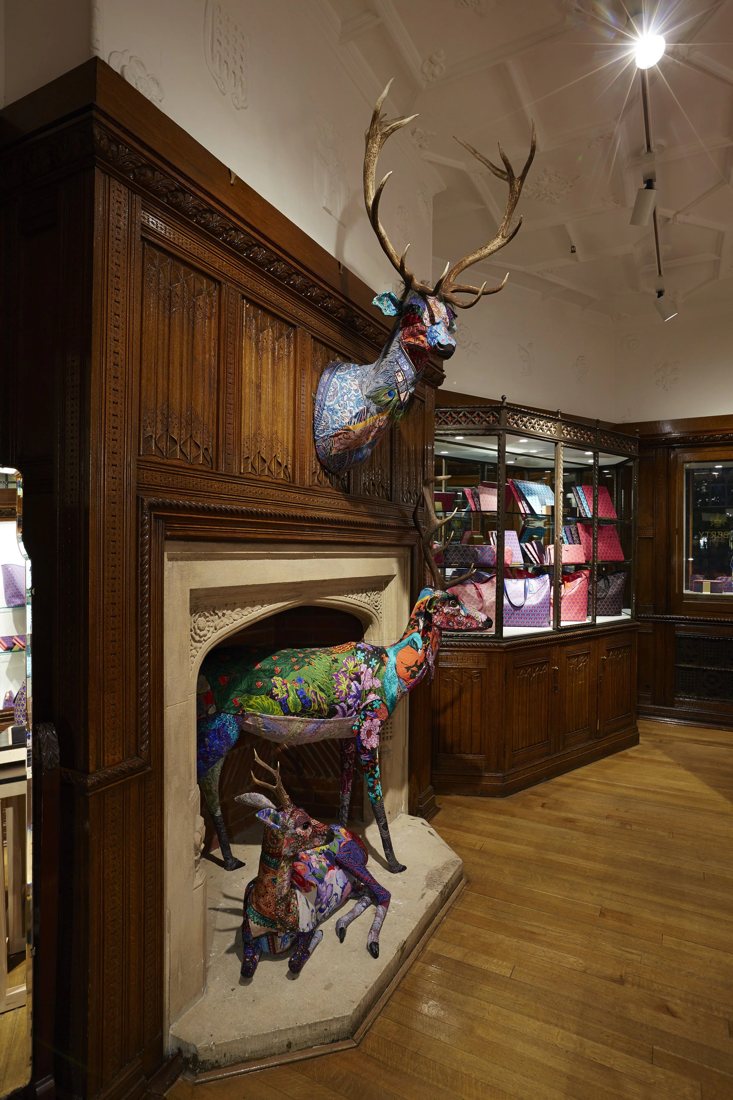 Colorful fabric art sculptures of three deer positioned on and around a fireplace with wooden paneling, in an interior space with wood flooring and glass display cases.