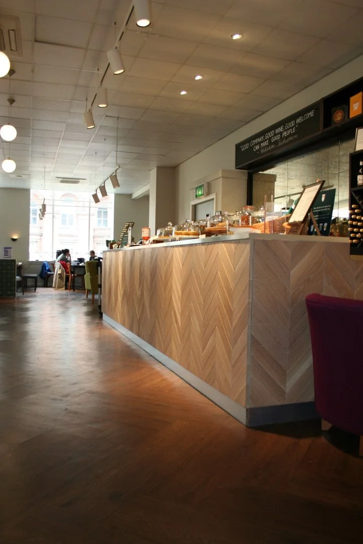A coffee shop counter with baked goods, a menu stand, and seating area in the background with people sitting near large windows.