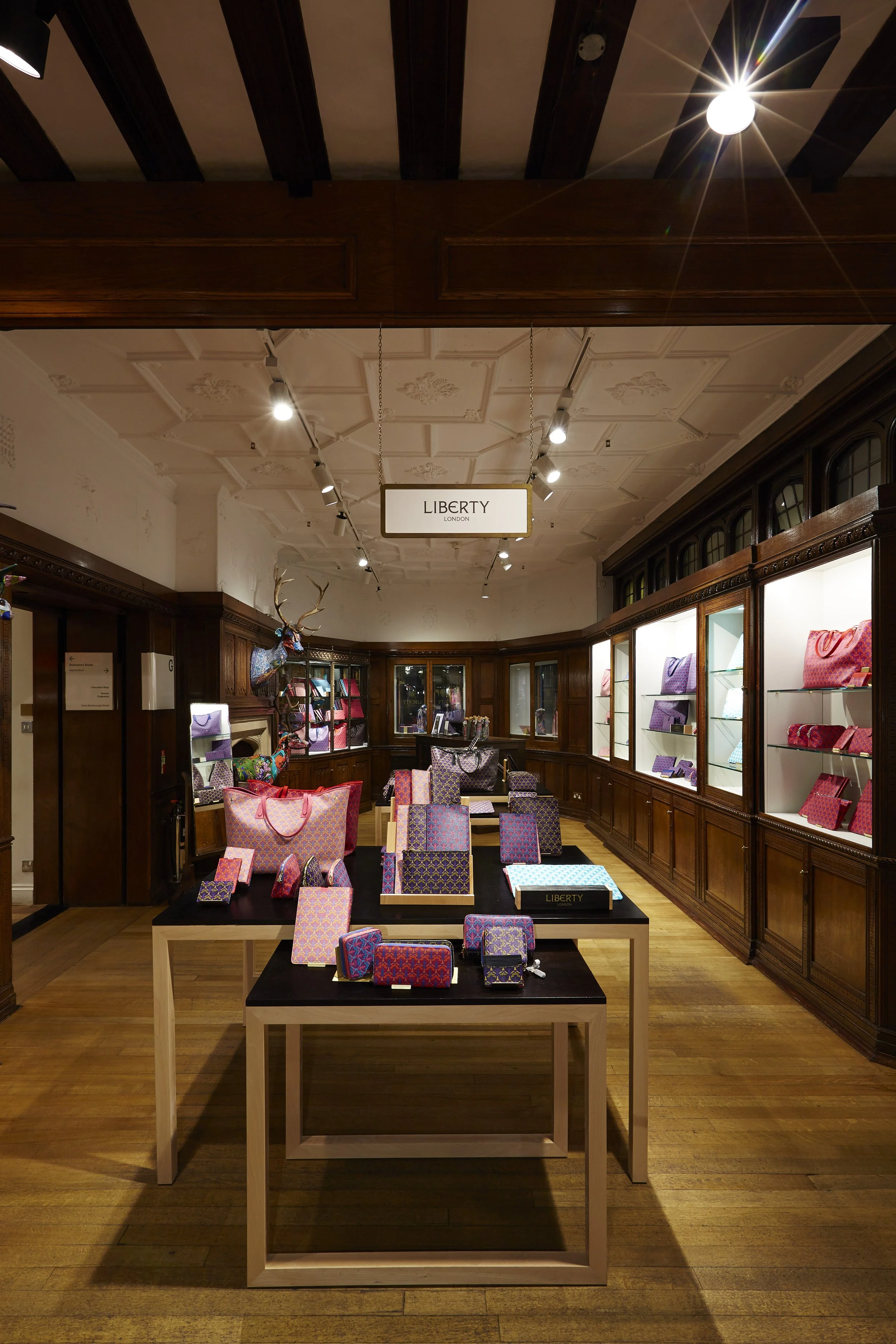 Interior of a luxury shop displaying colorful handbags on black tables and in glass display cabinets, with a sign hanging from the ceiling that says 'Liberty London'.