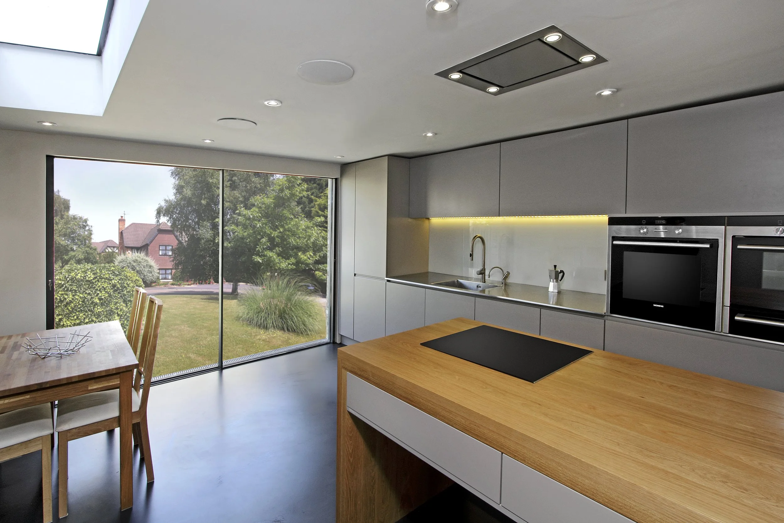 Modern kitchen with gray cabinets, wooden island, stainless steel sink, built-in oven, and large sliding glass door overlooking a grassy yard with trees and houses.