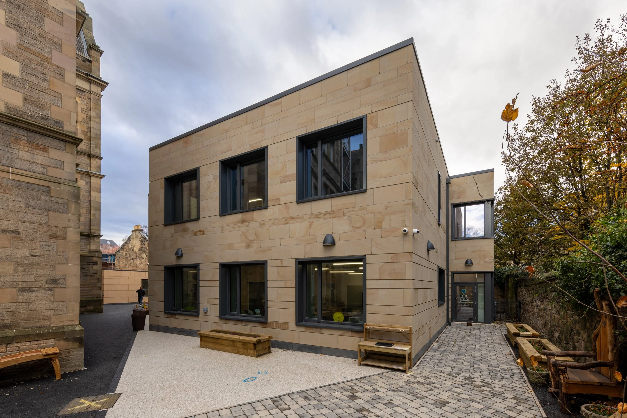 Modern multi-story building with beige stone exterior, black window frames, and outdoor seating, situated between older stone buildings and surrounded by trees and a cobblestone pathway.