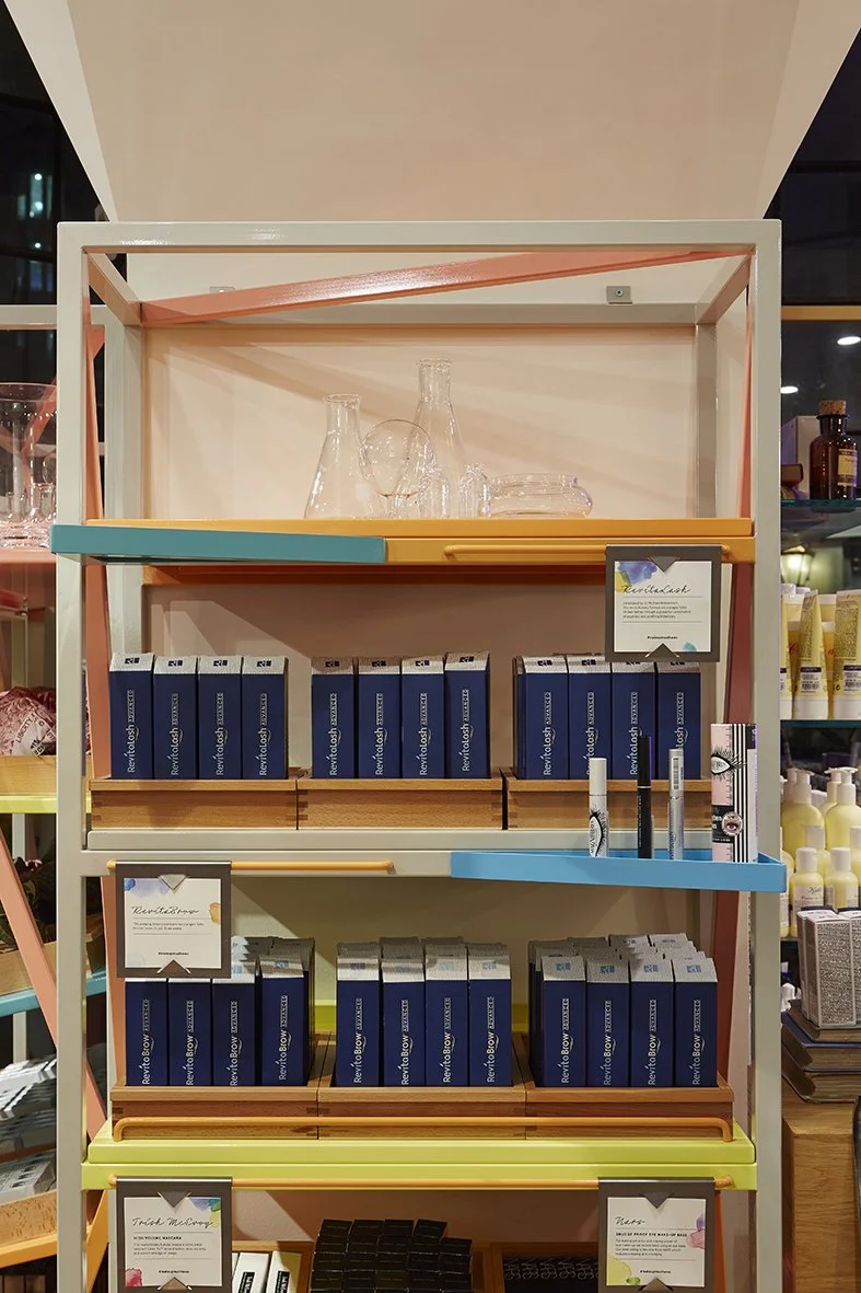 A retail display shelf with various skincare or cosmetic products, including boxed items labeled 'Revitalash' and pens, arranged on colorful shelves.