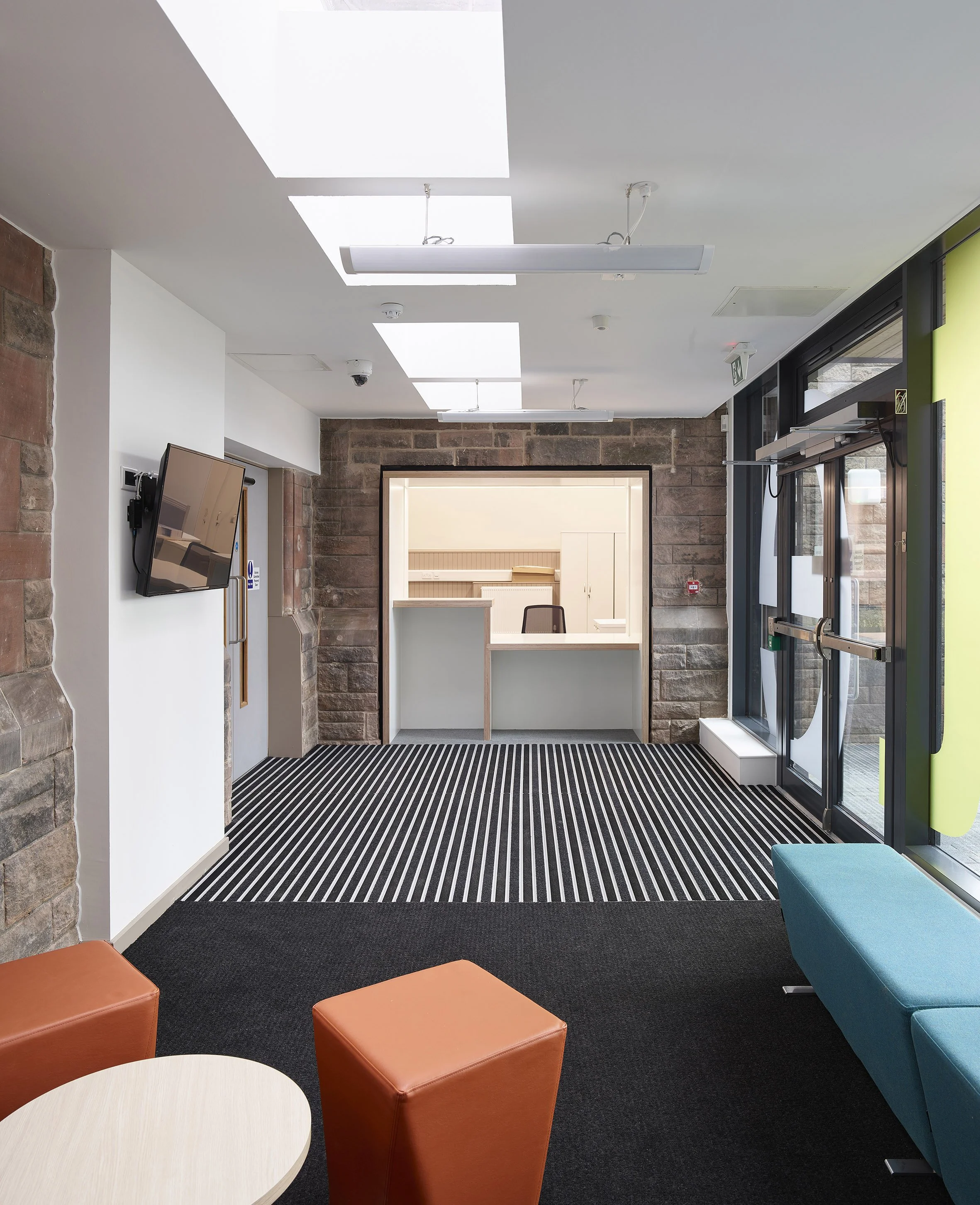 Interior view of a modern waiting area with a reception desk, striped carpet, orange and teal seating, a mounted TV, and large windows with glass doors.