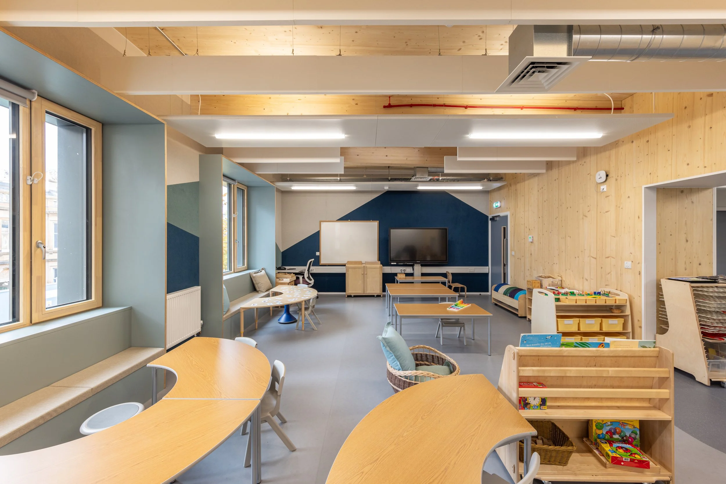 Empty classroom or playroom with wooden furniture, tables, chairs, educational materials, and a large screen on the wall.