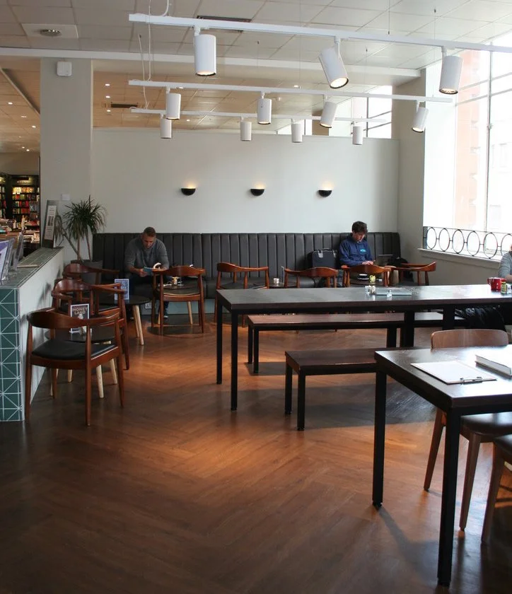 Inside a bright, modern café with wooden floors, black tables, and wooden chairs. Two customers sit at separate tables near a white wall with black wall sconces, and large windows let in natural light.