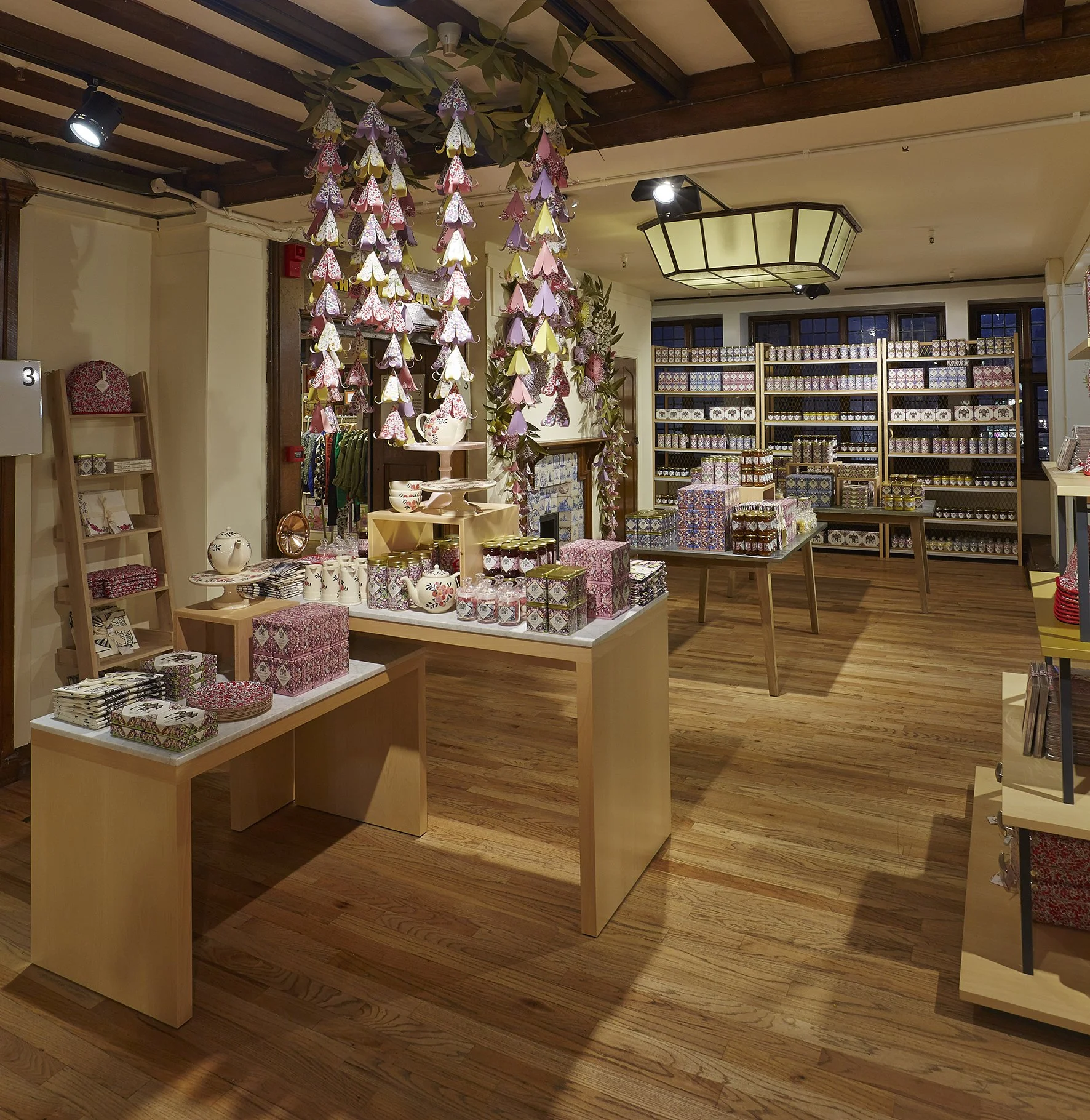 A retail store interior decorated with pink, purple, and yellow paper butterfly garlands hanging from the ceiling. The store has wooden floors, shelves stocked with patterned boxes and ceramics, and tables displaying assorted products.