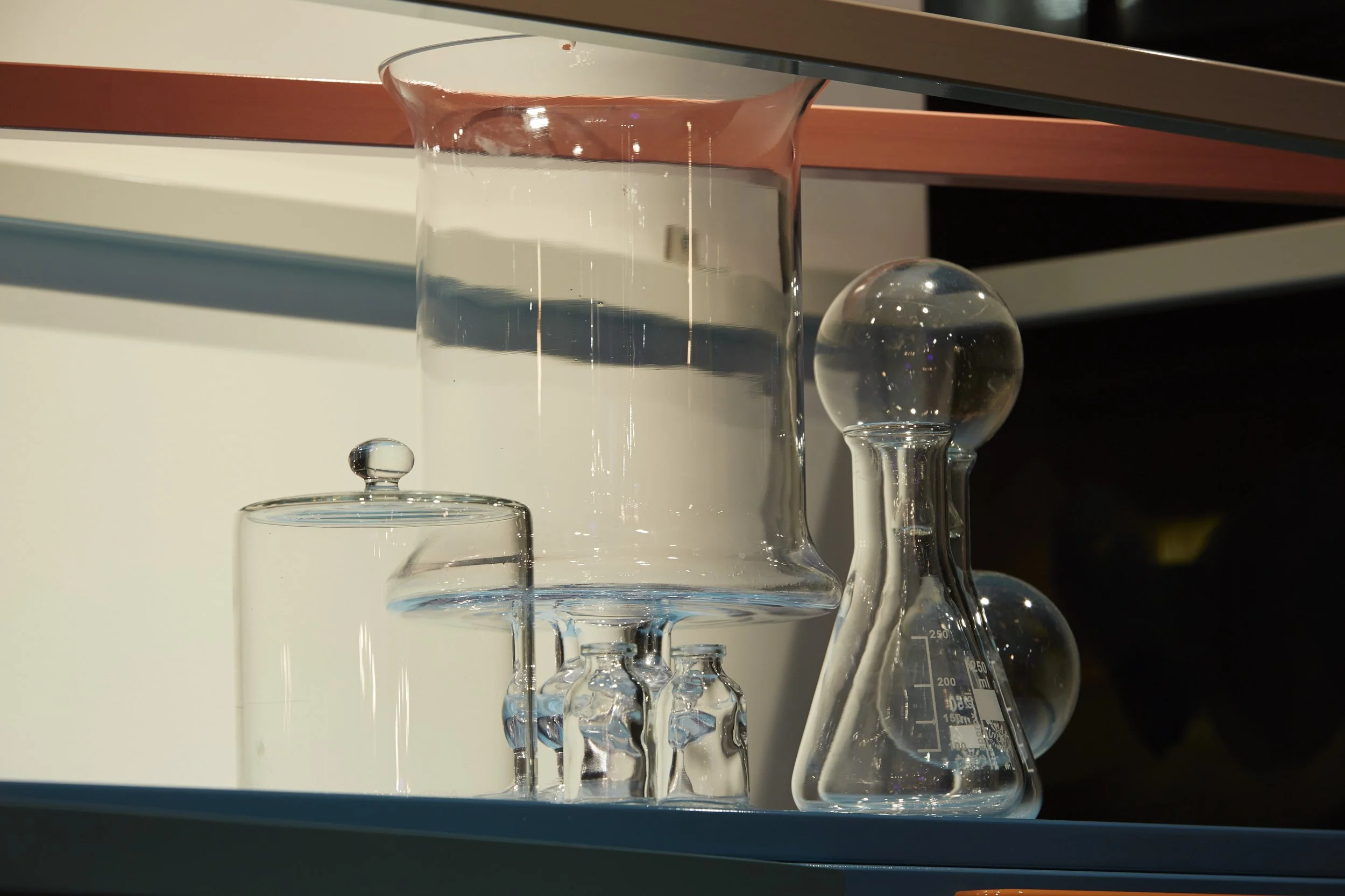 Various laboratory glassware, including a large beaker filled with water, a smaller beaker, a conical flask with measurement markings, and several round-bottom flasks, all placed on a shelf.