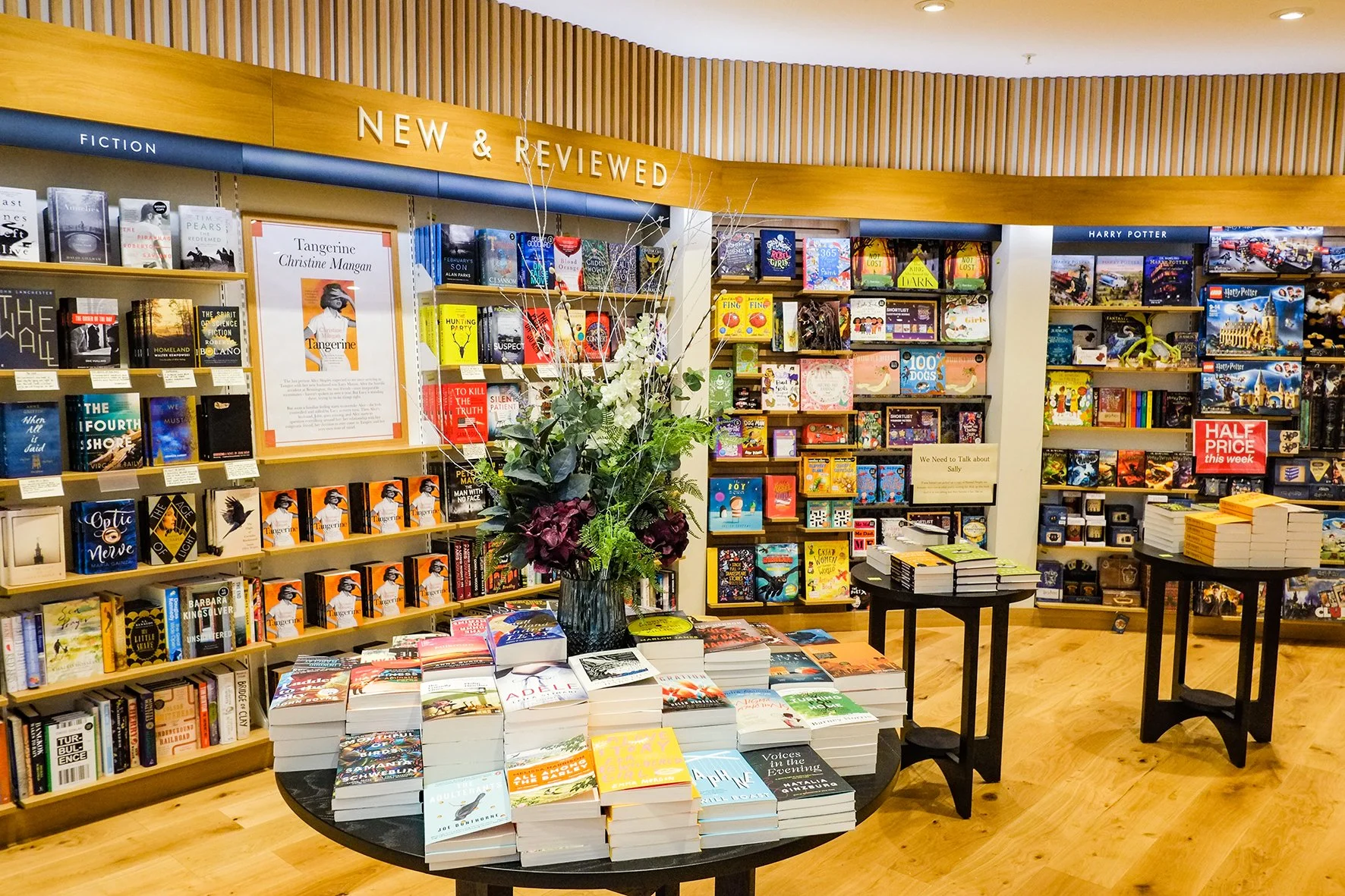 Bookshelves with new and reviewed books, display tables with stacks of books, and a large floral arrangement in the center inside a bookstore.