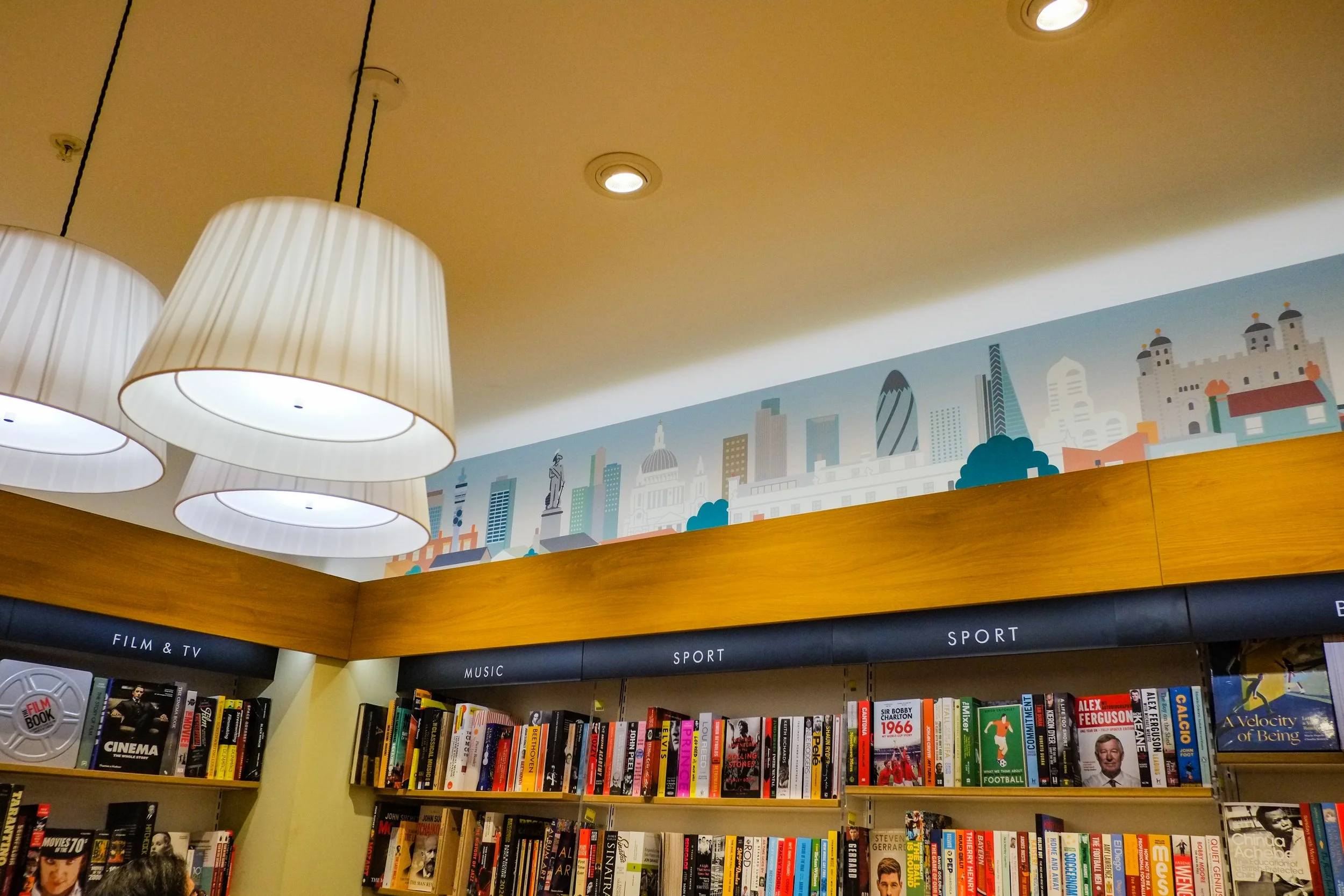 Bookshelves in a bookstore with section labels 'Film & TV,' 'Music,' and 'Sport,' a cityscape mural on the wall, and large hanging lamps.