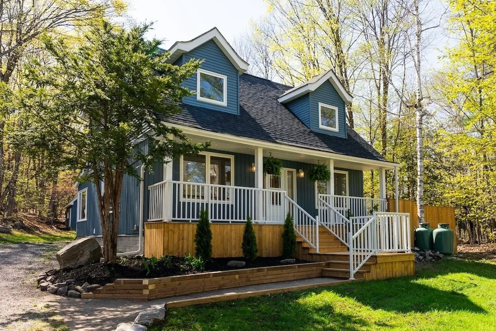 Muskoka cottage exterior with covered front porch, blue siding, and landscaped yard surrounded by trees.
