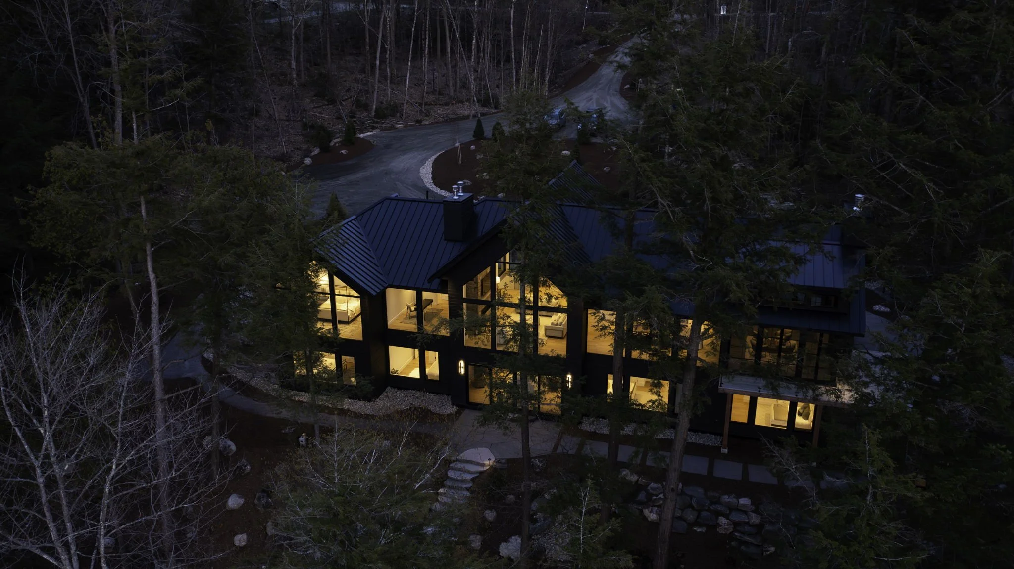 Aerial night view of a luxury Muskoka cottage surrounded by trees with warm interior lighting.