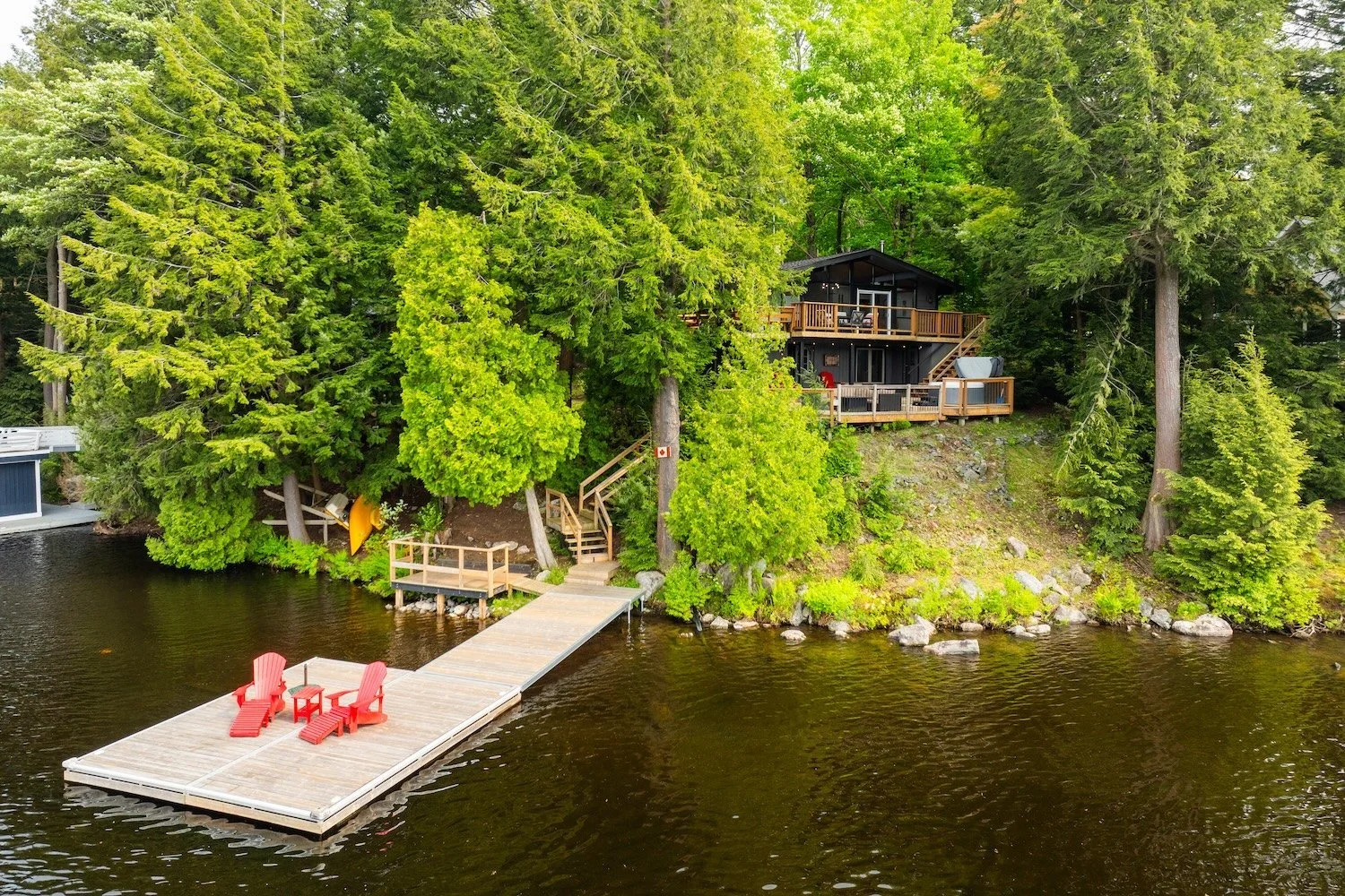 Lakeside cottage surrounded by trees with private dock and red Muskoka chairs on the water.