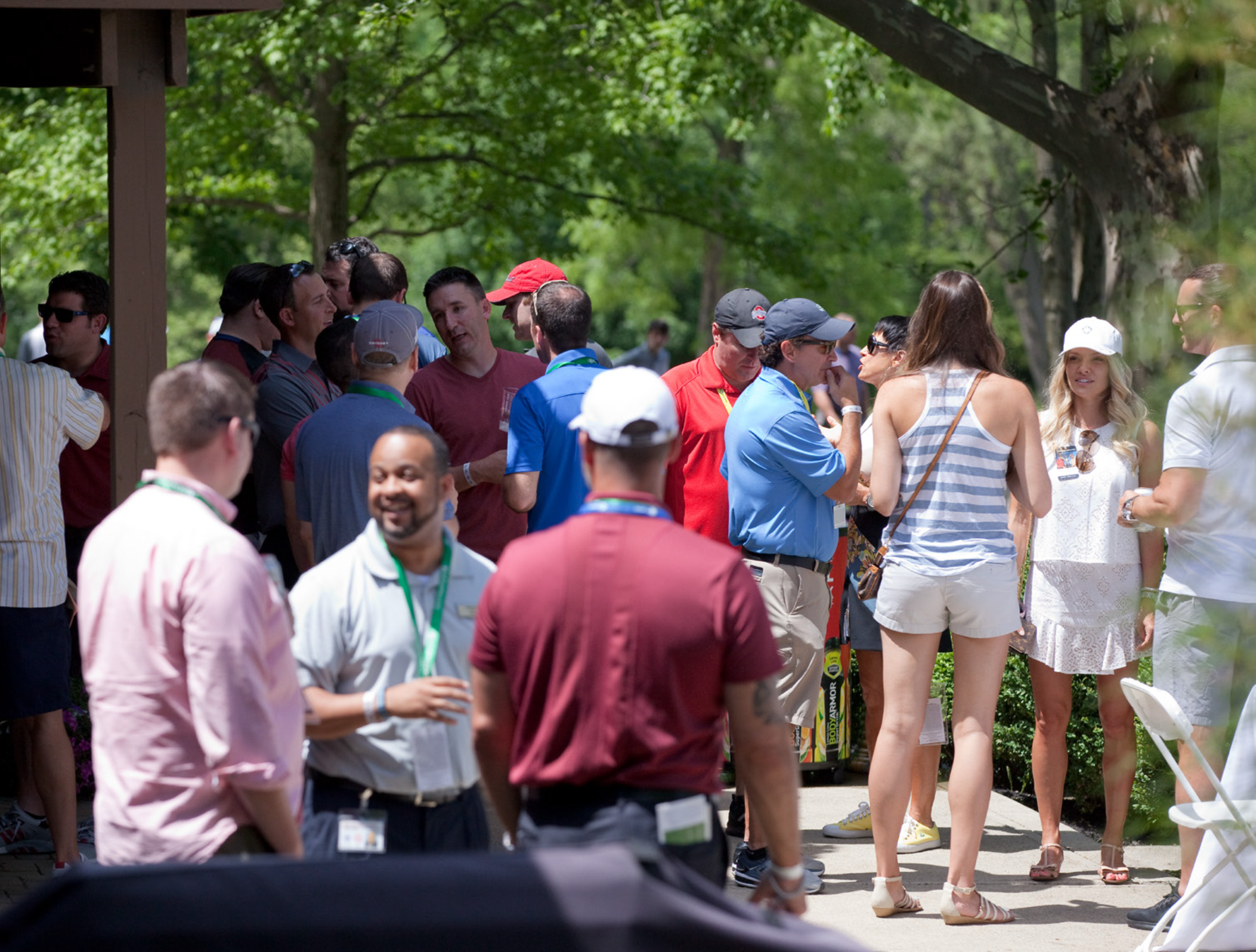Group of people socializing outdoors on a sunny day, some wearing sunglasses and hats, with trees and greenery in the background.