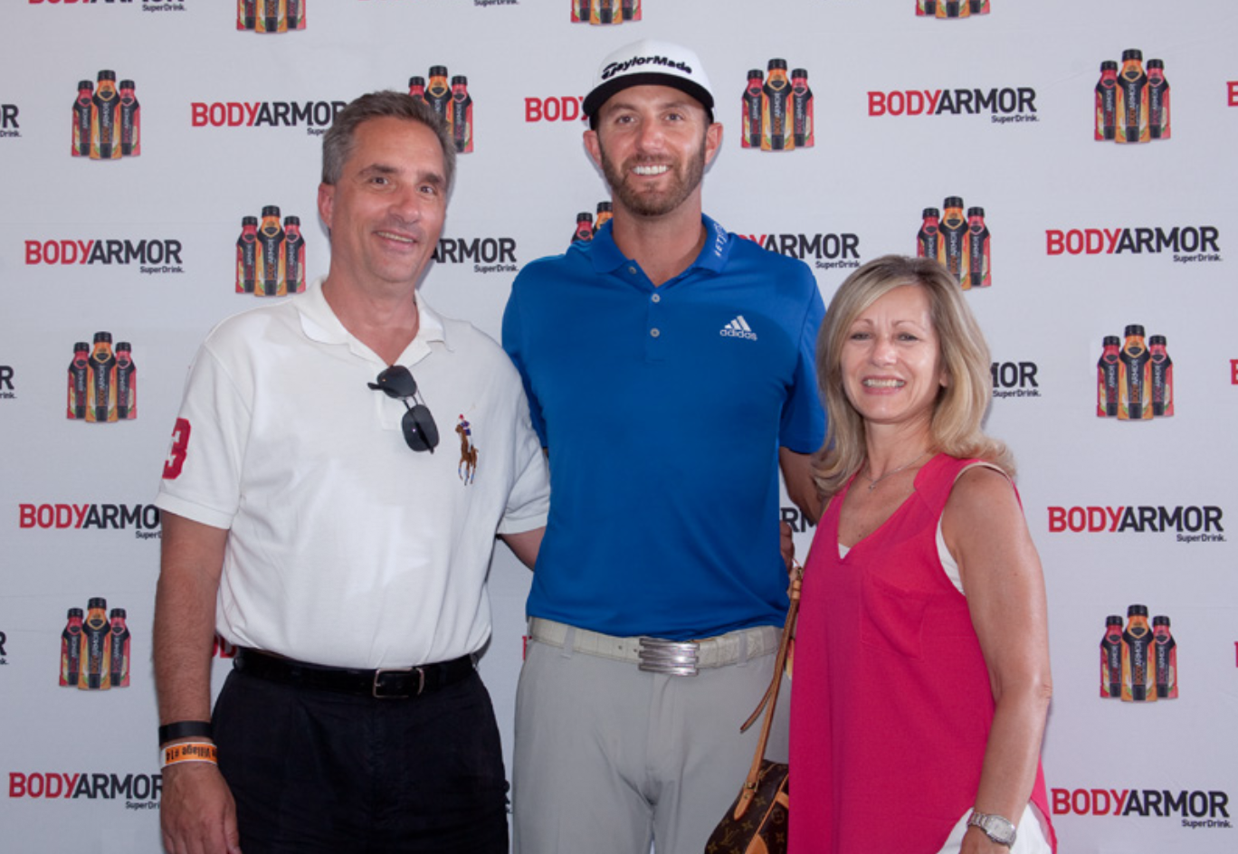 Three people at a promotional event standing in front of a backdrop with the logo BODARMOR and images of bottled drinks. The man on the left is wearing a white polo shirt with sunglasses hanging on his collar. The man in the middle is wearing a blue polo shirt and cap, and the woman on the right is wearing a pink sleeveless top with a wristwatch.
