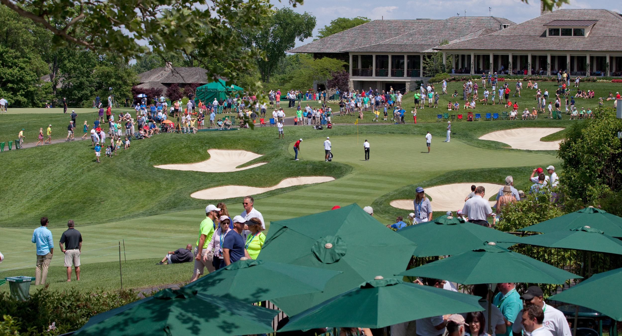 A golf course scene with multiple sand traps, a putting green, and numerous people, including spectators and players, enjoying a sunny day. Some people are sitting, walking, or watching the game, with a clubhouse or large building in the background and green umbrellas providing shade in the foreground.