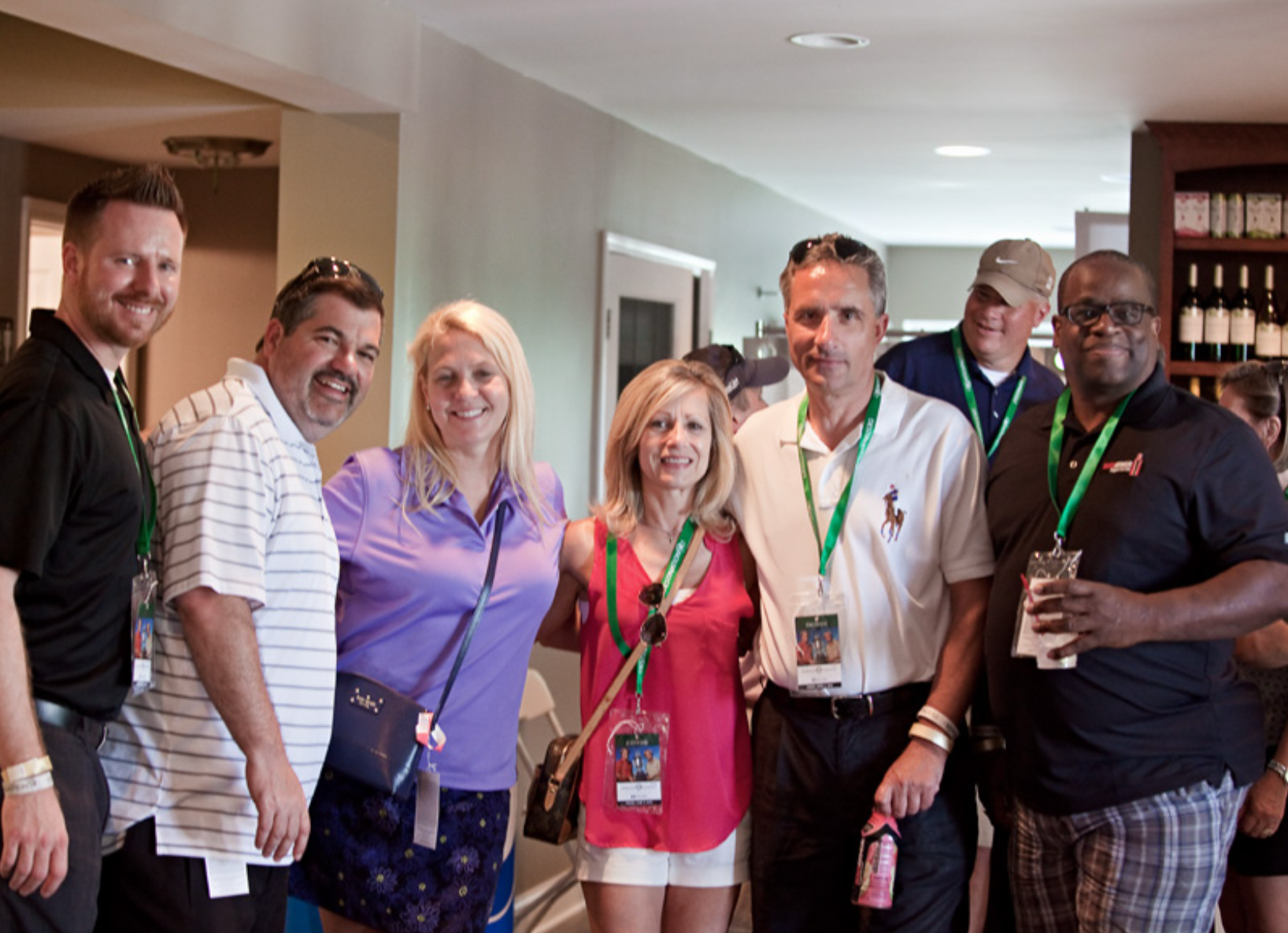 Group of seven adults standing inside a building, smiling, wearing casual clothes, with some holding drinks and conference badges on green lanyards.