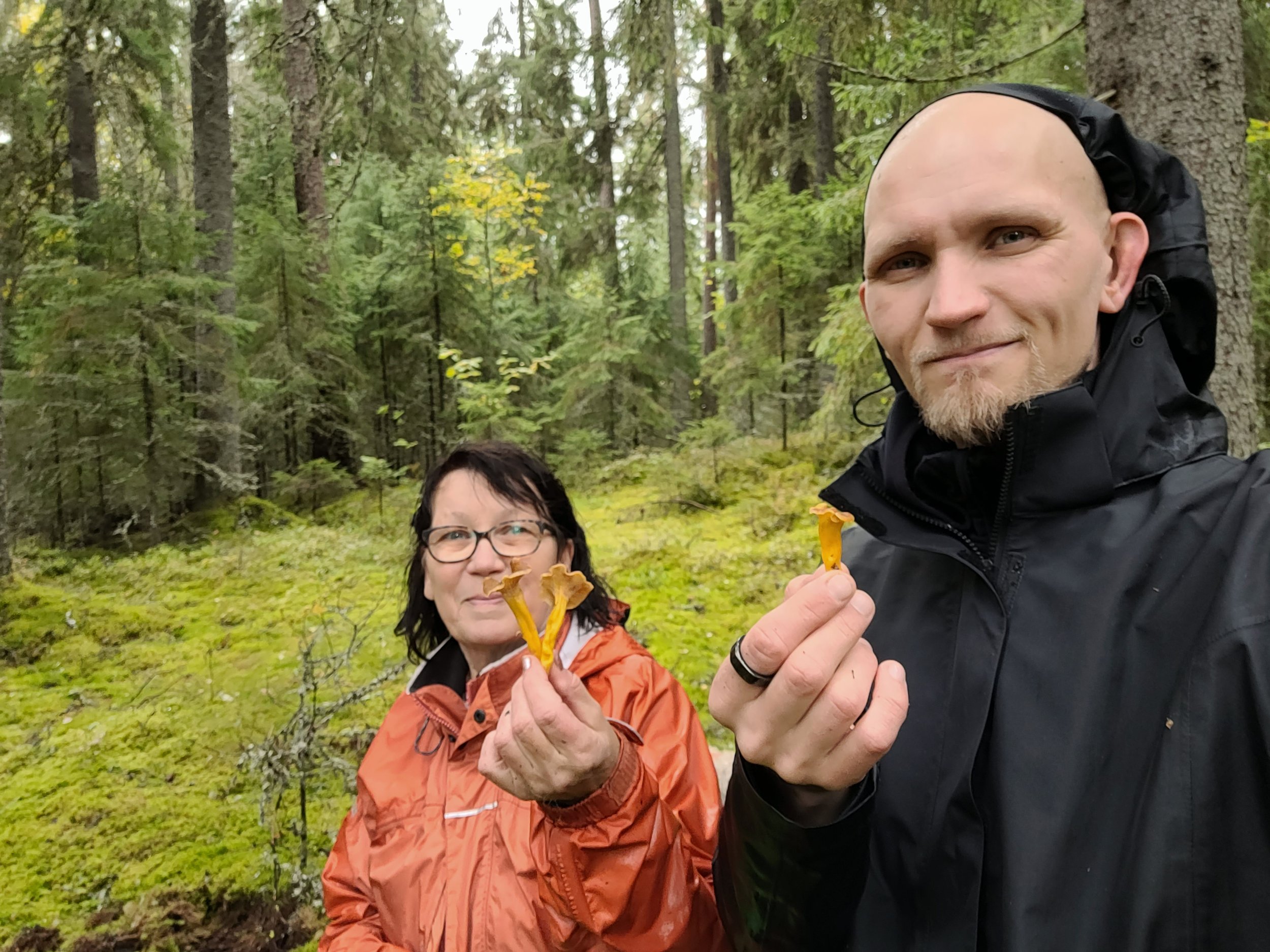 Kuvassa kaksi ihmistä metsässä, molemmat pukeutuneet ulkoiluvaatteisiin ja näyttävät nauttivan retkestä. Molemmat pitävät kädessään kanttarelleja.