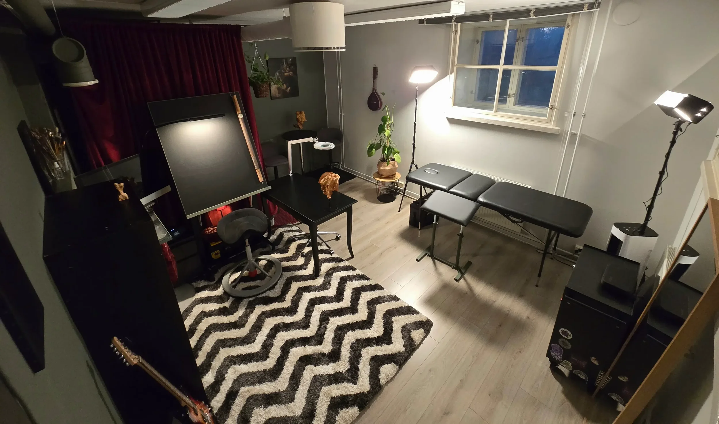 Kuva kotoisasta taide- ja tatuointistudiosta, jossa on musiikki- ja työskentelyvälineitä, kuten kitara, studio-valot, pöytä, tuoli. Huoneessa on matto, kasvi ja ikkuna.