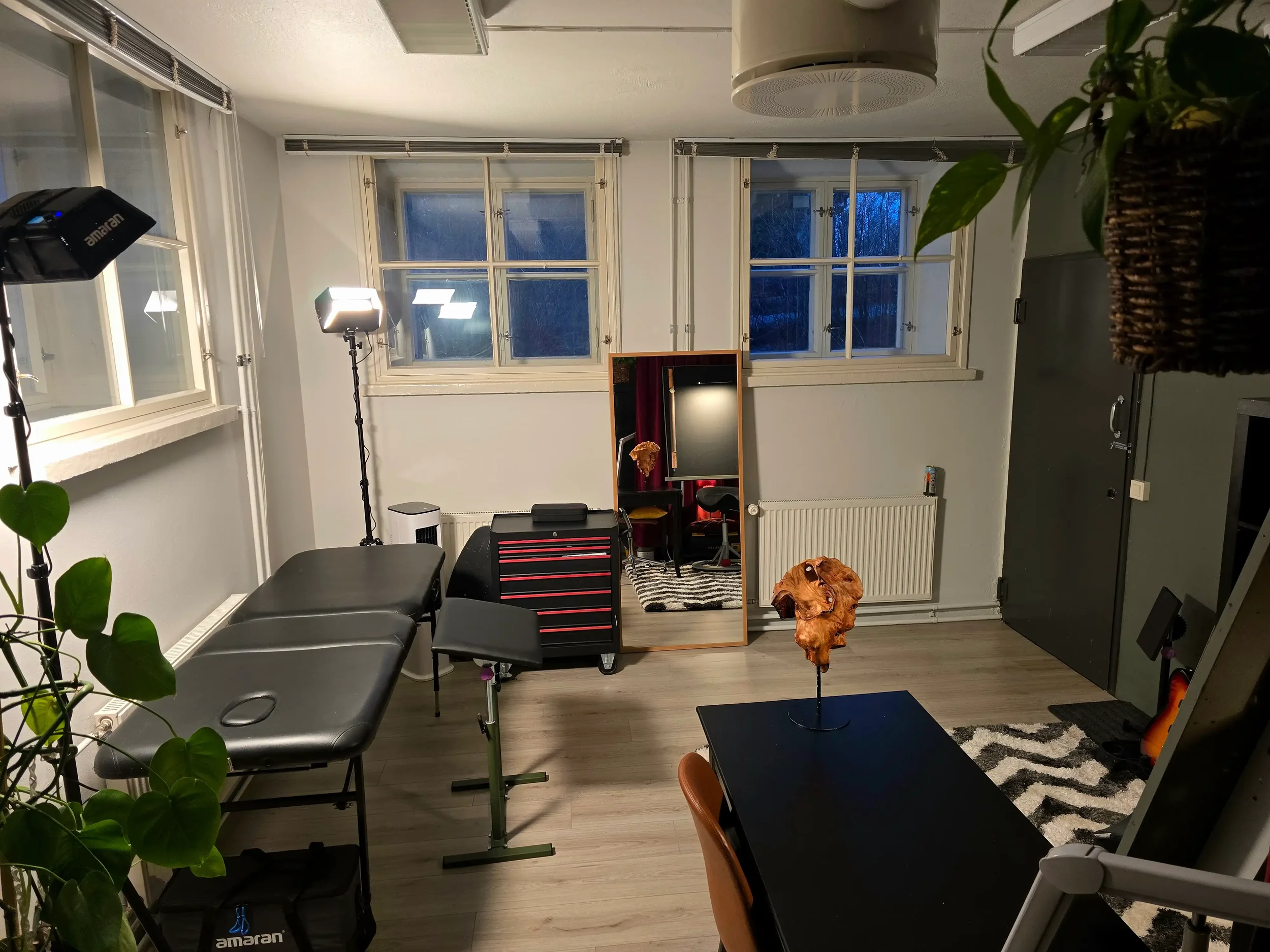 Valoisa taide-ja tatuointistudio jossa on hierontapöytä, peili, valo- ja kameravaloja ja pieni kirjoituspöytä, sekä kolme ikkunaa.