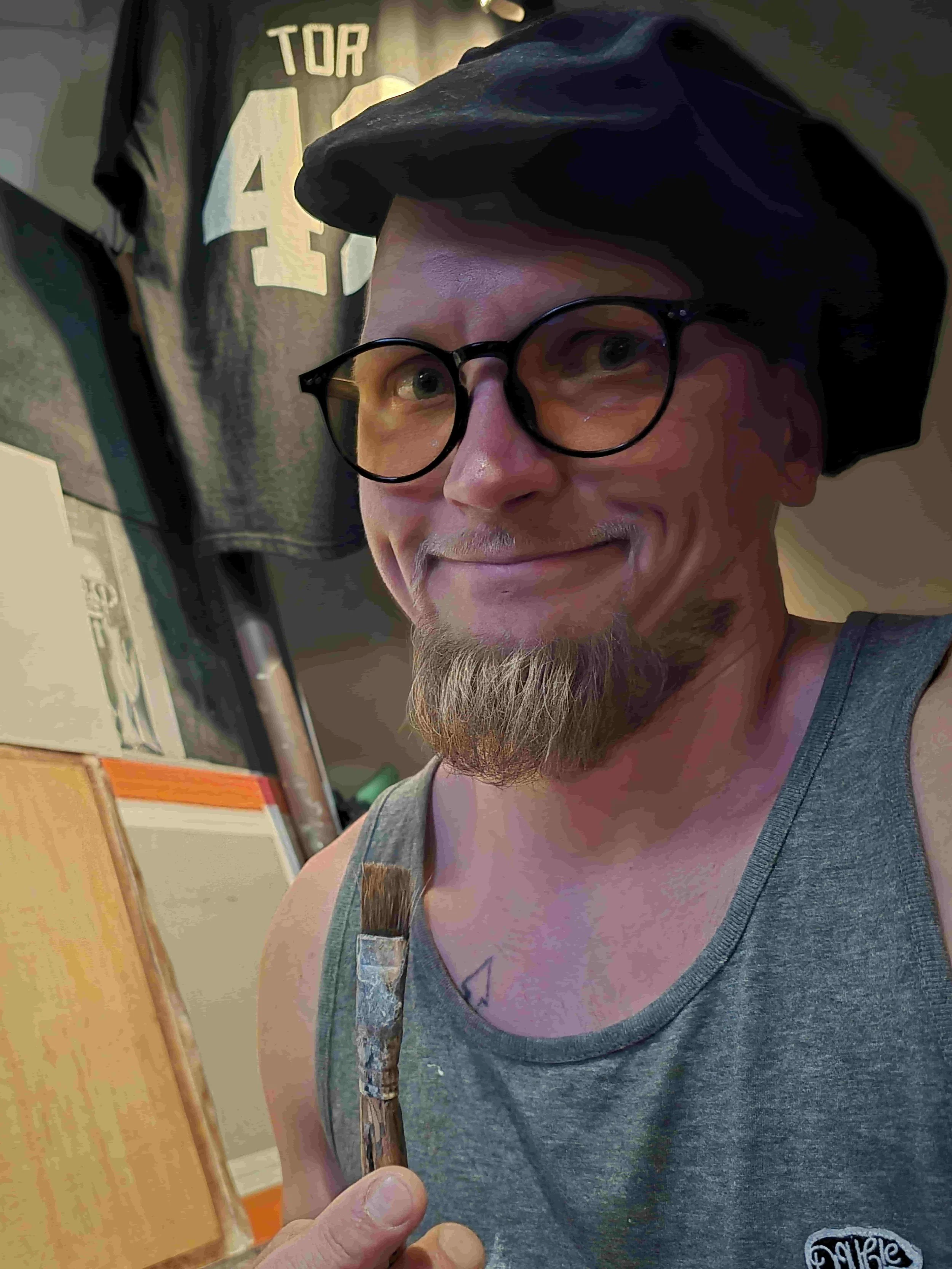Kuvassa suuri taiteilija, jolla on pyöreät silmälasit ja musta baskeri, katsoo kameraan ja hymyilee vastustamattomasti.