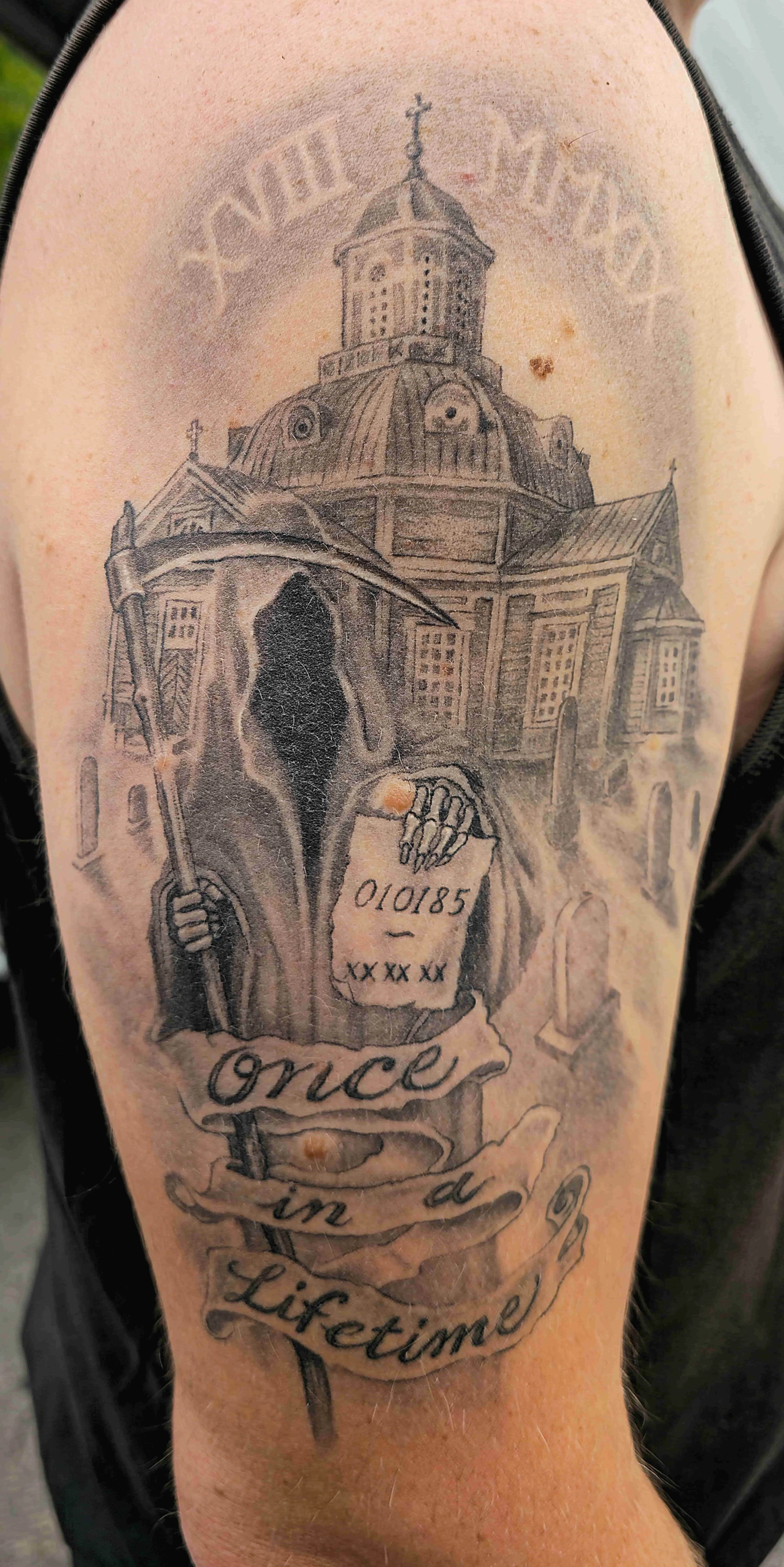 Mustavalkoinen tatuointi, joka kuvaa hautausmaalta nousevaa kuoleman hahmoa, hautakiviä ja kirkko, tekstit "Once in a Lifetime" ja paremmalla puolella myös "010185".