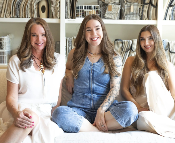 Three women sitting closely together on a beige couch, smiling, in a room with shelves of fabric or upholstery samples in the background.