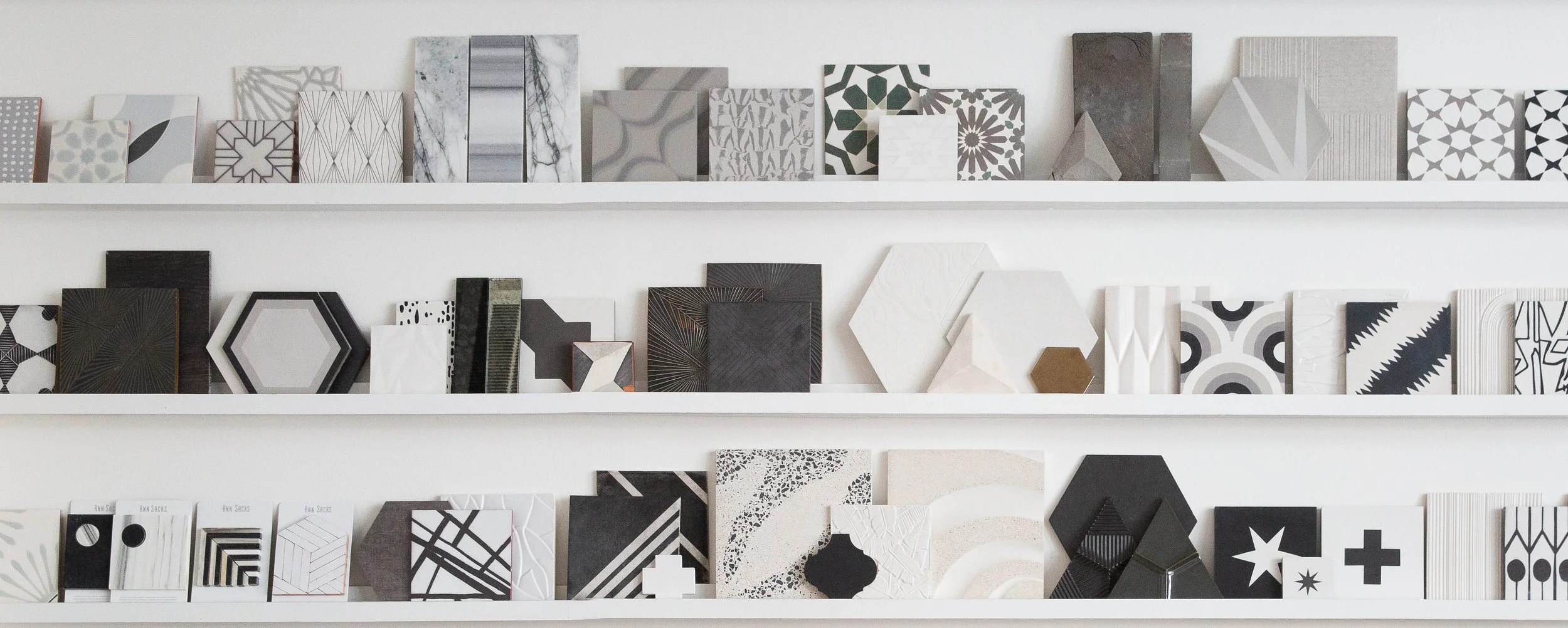 Display of various tiles on white shelves, featuring geometric, abstract, and patterned designs in black, white, gray, and muted tones.