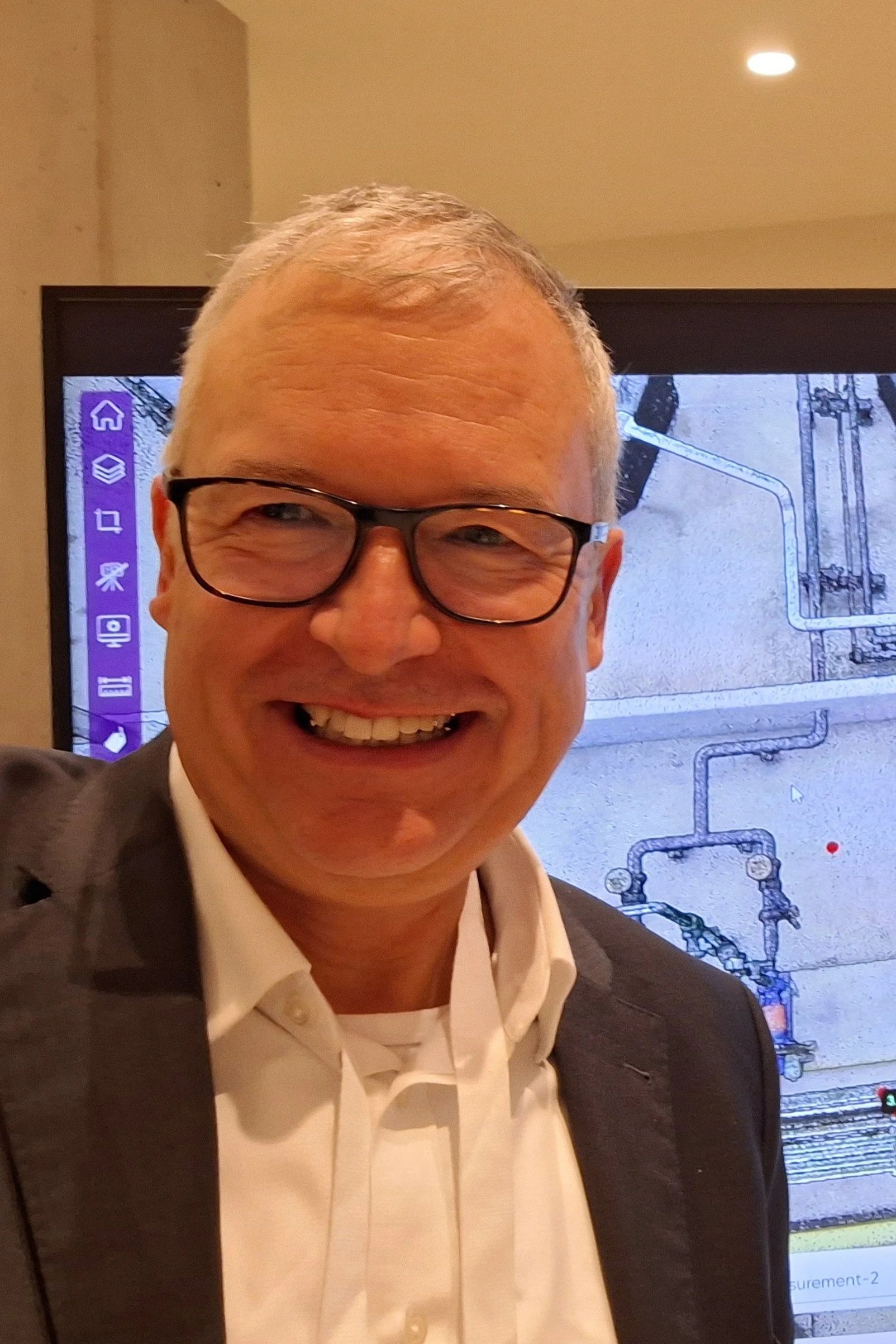 Close-up of a smiling man with glasses, wearing a white shirt and a dark blazer, standing in front of a screen displaying a schematic diagram.