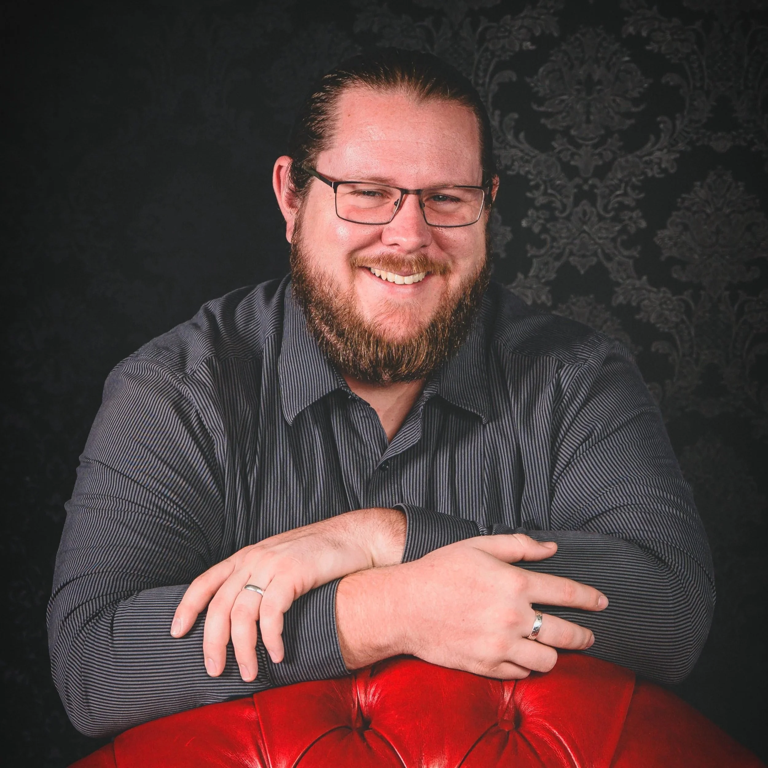 A smiling man with glasses and a beard, wearing a dark striped shirt, leaning on a red leather chair with a patterned black background.