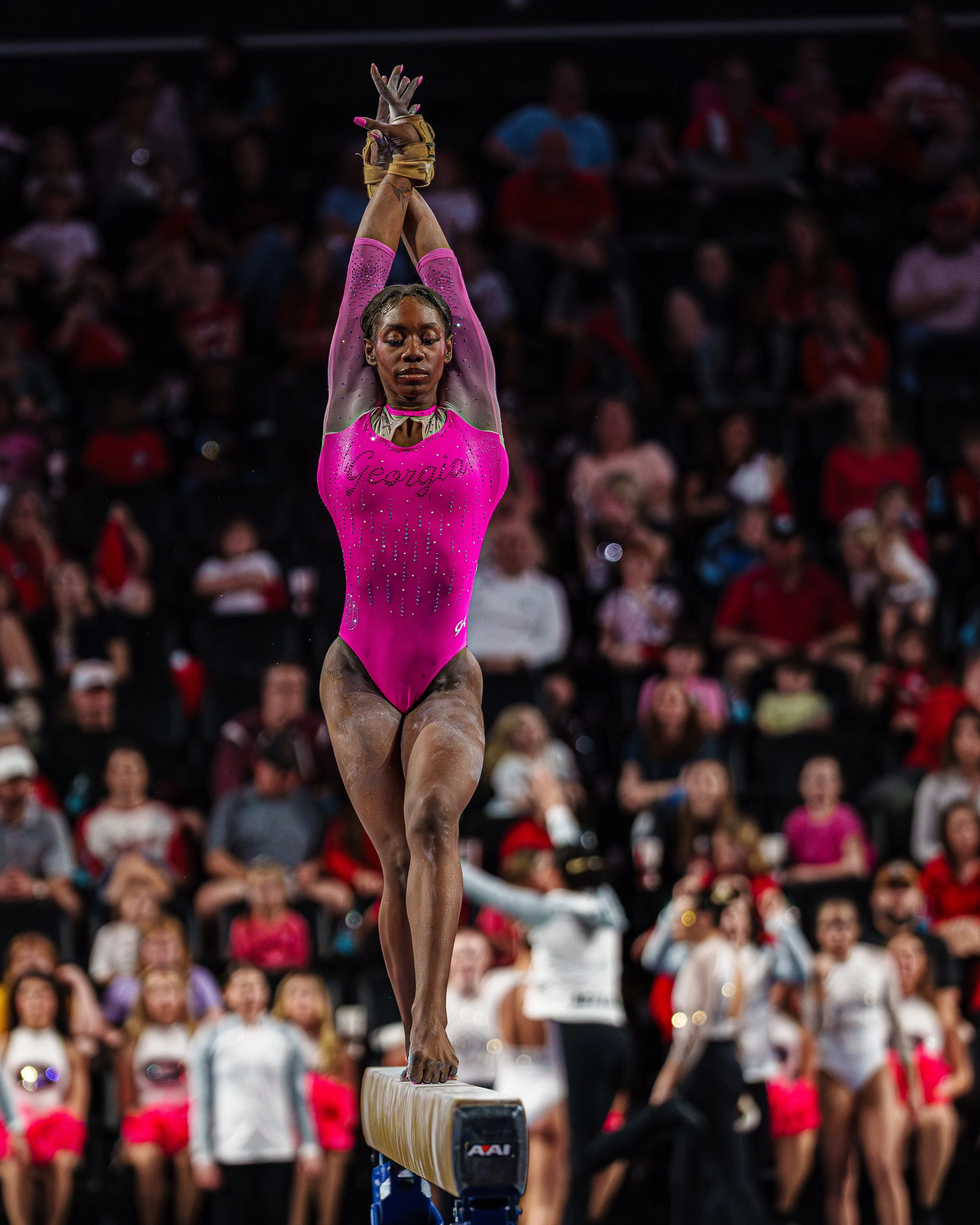 A female gymnast in a pink leotard balancing on a beam during a gymnastics competition, with an audience watching in the background.