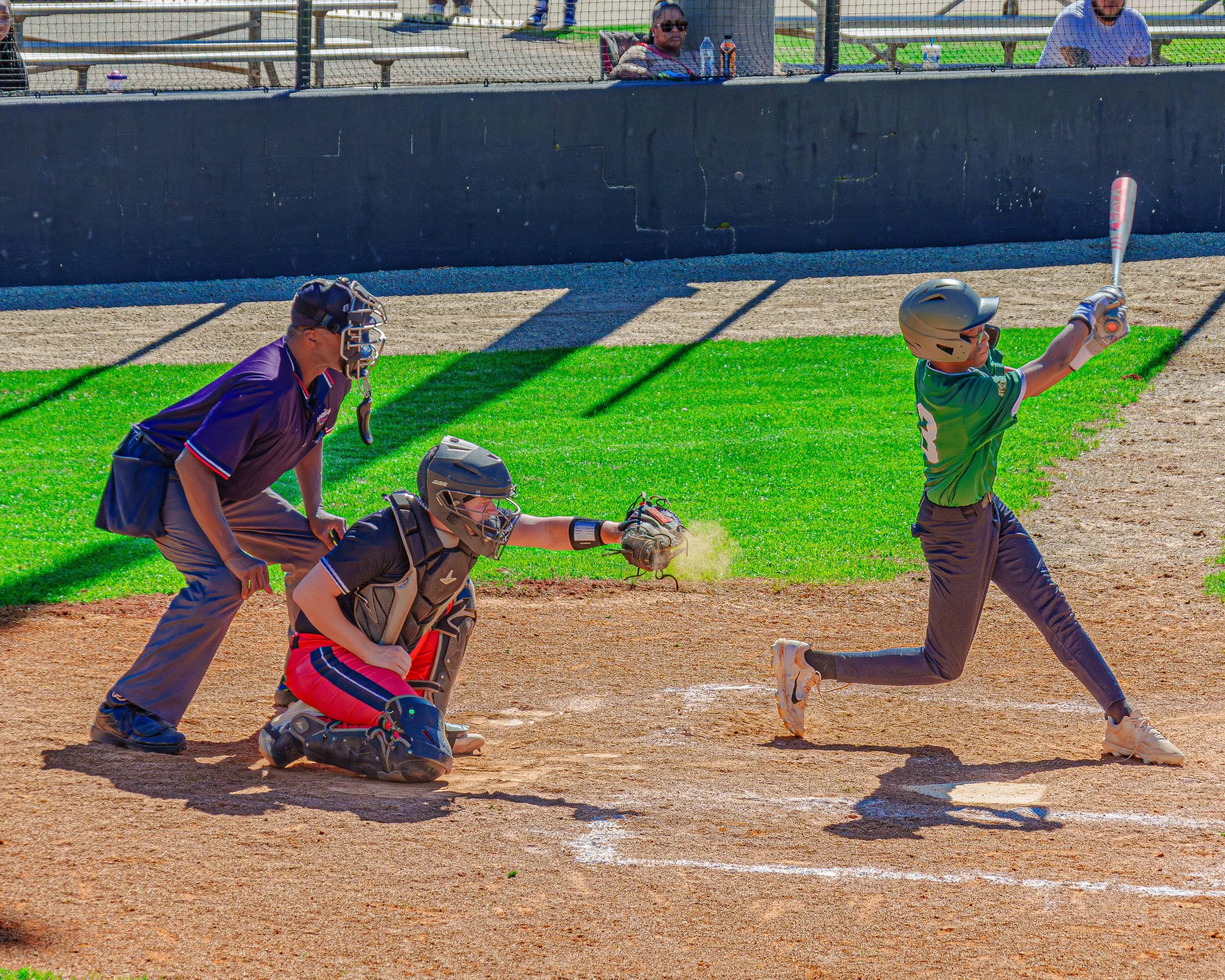 A baseball player in a green jersey and gray helmet swinging a bat, with the ball about to be hit. The catcher and umpire behind home plate are in position, and spectators are watching from behind the fence.