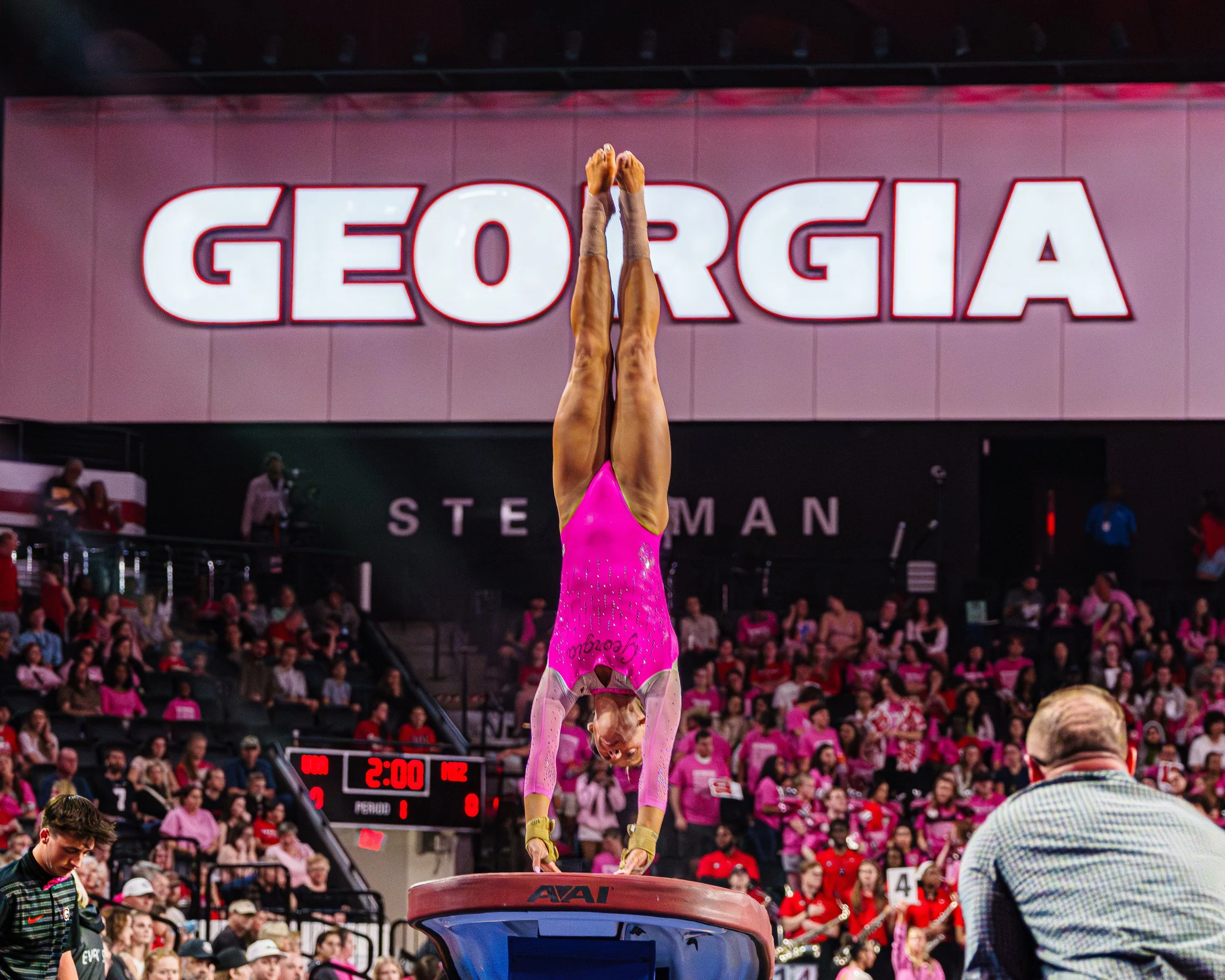 Gymnast performing a handstand on the vault at a competition with a large Georgia banner in the background and an audience in pink.