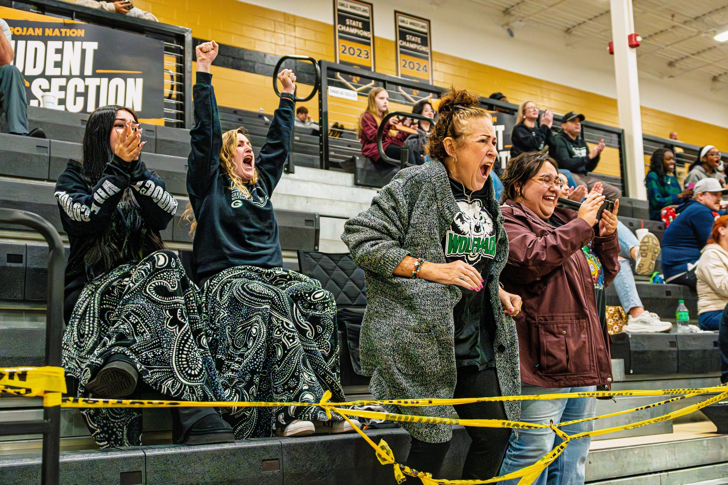 Group of women reacting excitedly, cheering and smiling at a sports event in a gymnasium, with some taking photos and others clapping.