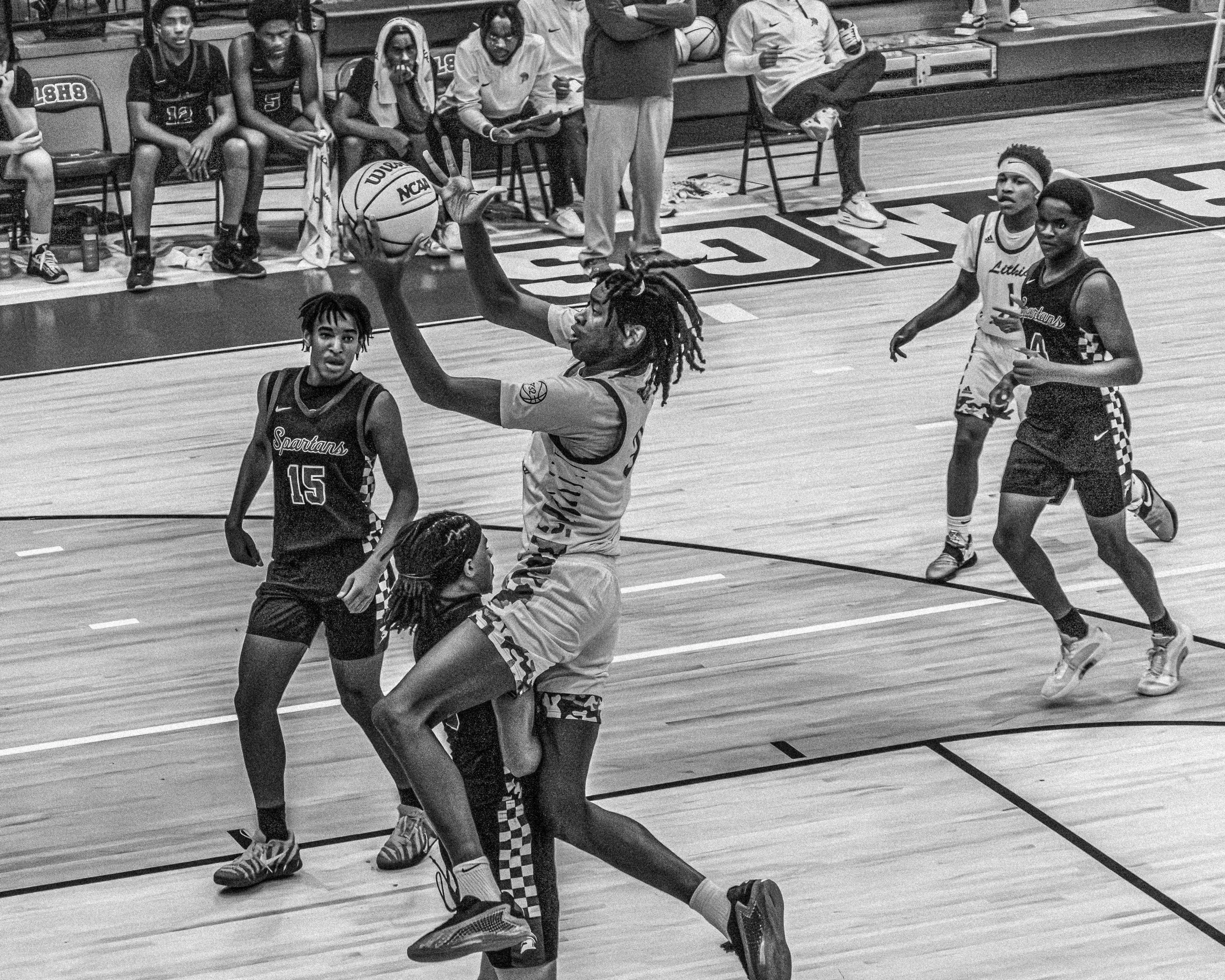 A basketball player jumping to shoot or pass the ball during a game, surrounded by opponents on the court with players sitting on the bench in the background.