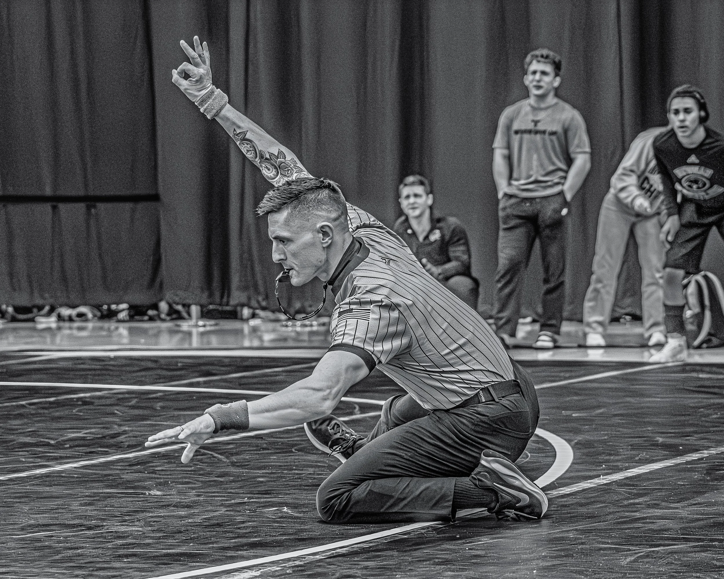 A wrestling referee kneeling on a wrestling mat, holding a whistle in his mouth and reaching forward with his right hand. In the background, three spectators and two wrestlers are watching a match.