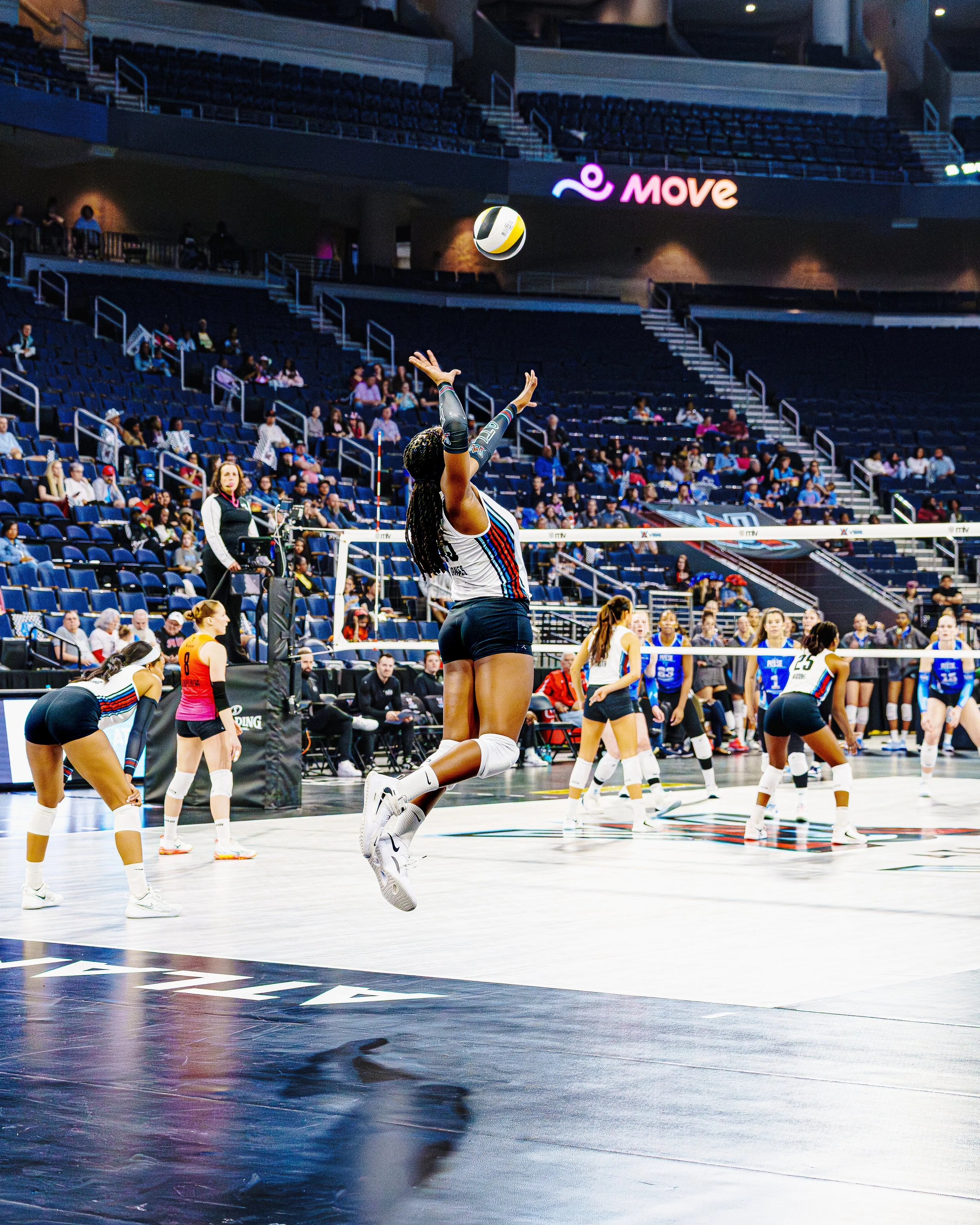 A volleyball player in a white and blue uniform jumps to hit the ball during a game in an indoor arena with spectators.