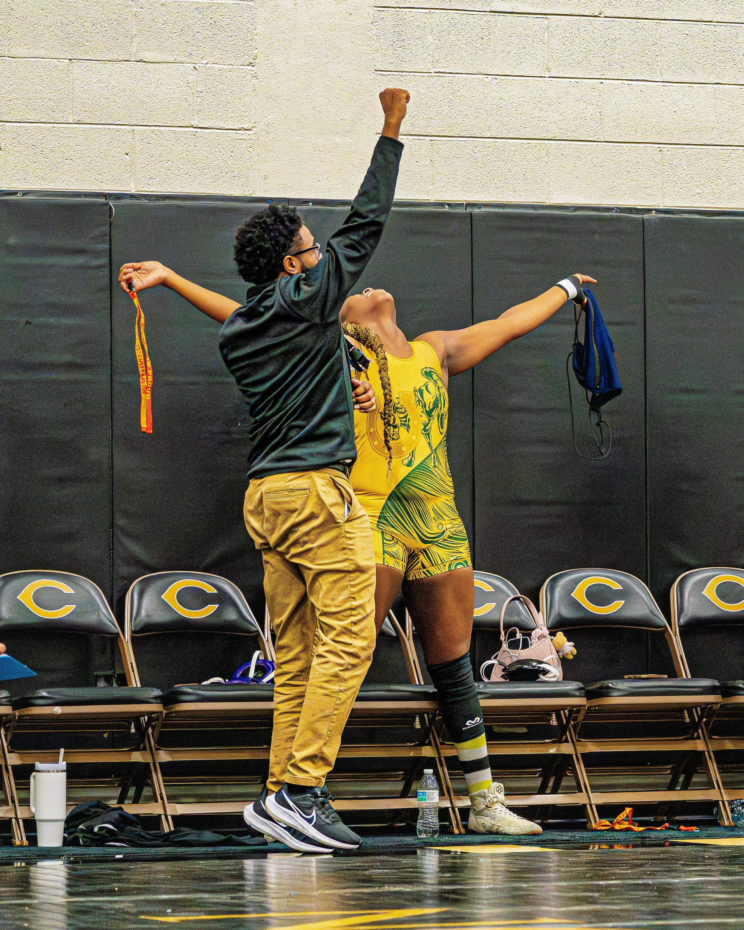 A female athlete in a yellow sports uniform celebrating with her hands raised in a gymnasium, while a person next to her appears to be cheering or encouraging her.