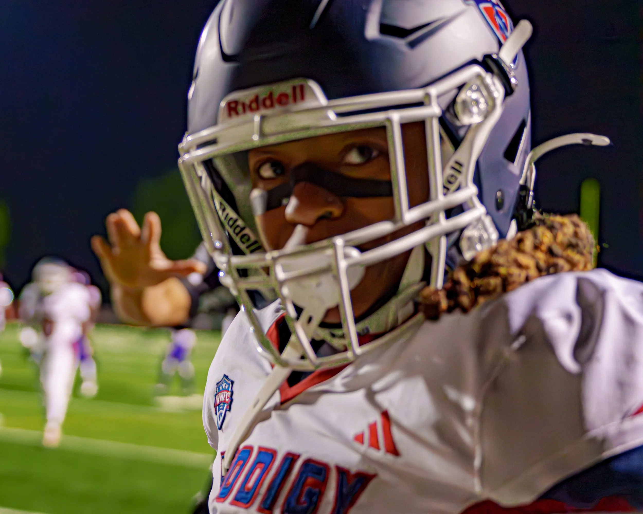 Close-up of a football player in a helmet with a face mask, wearing a white jersey with red and blue accents, on a football field at night with other players in the background.