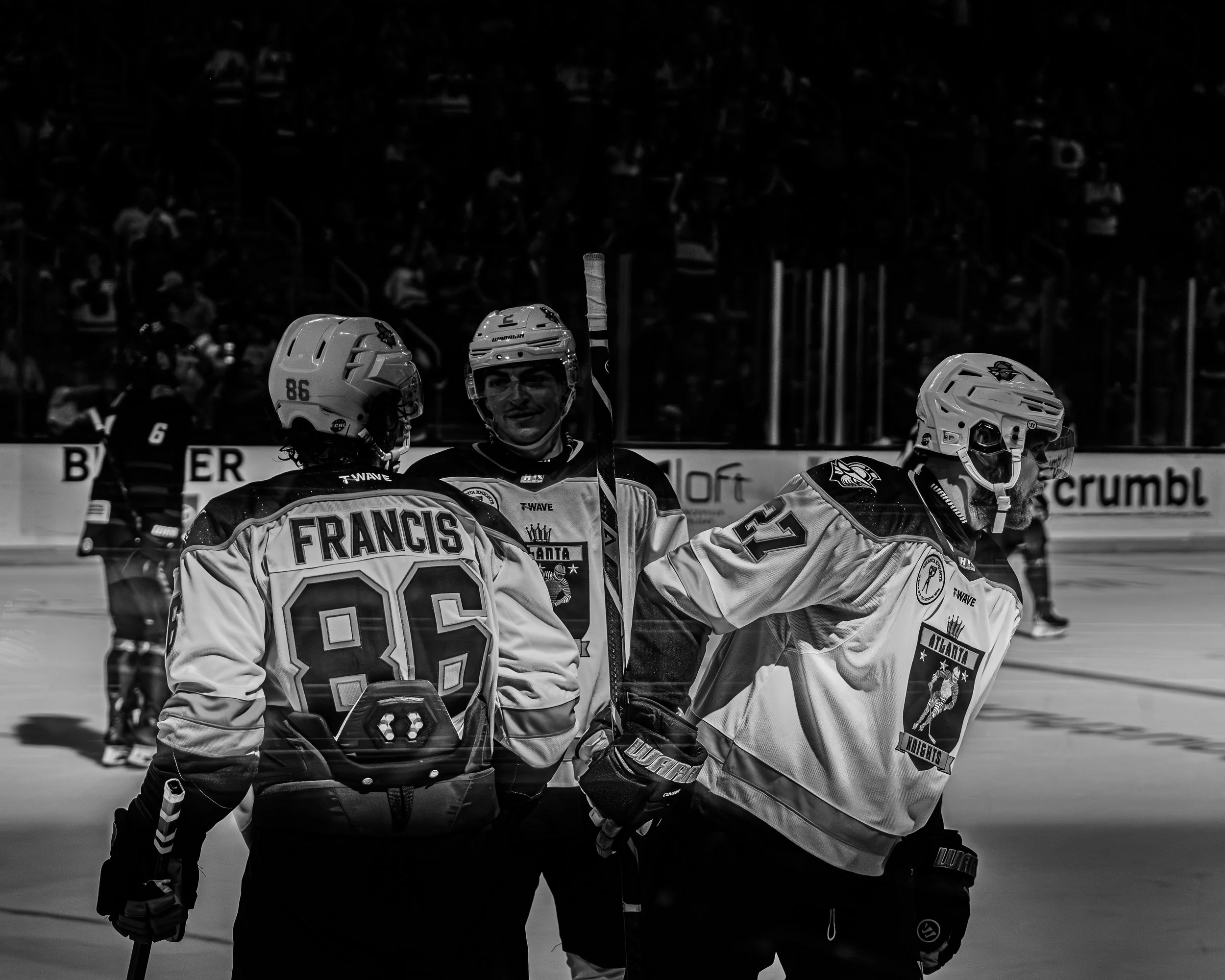 Three ice hockey players on the rink, two facing away from the camera and one facing forward, wearing jerseys with the Atlanta Knights logo.