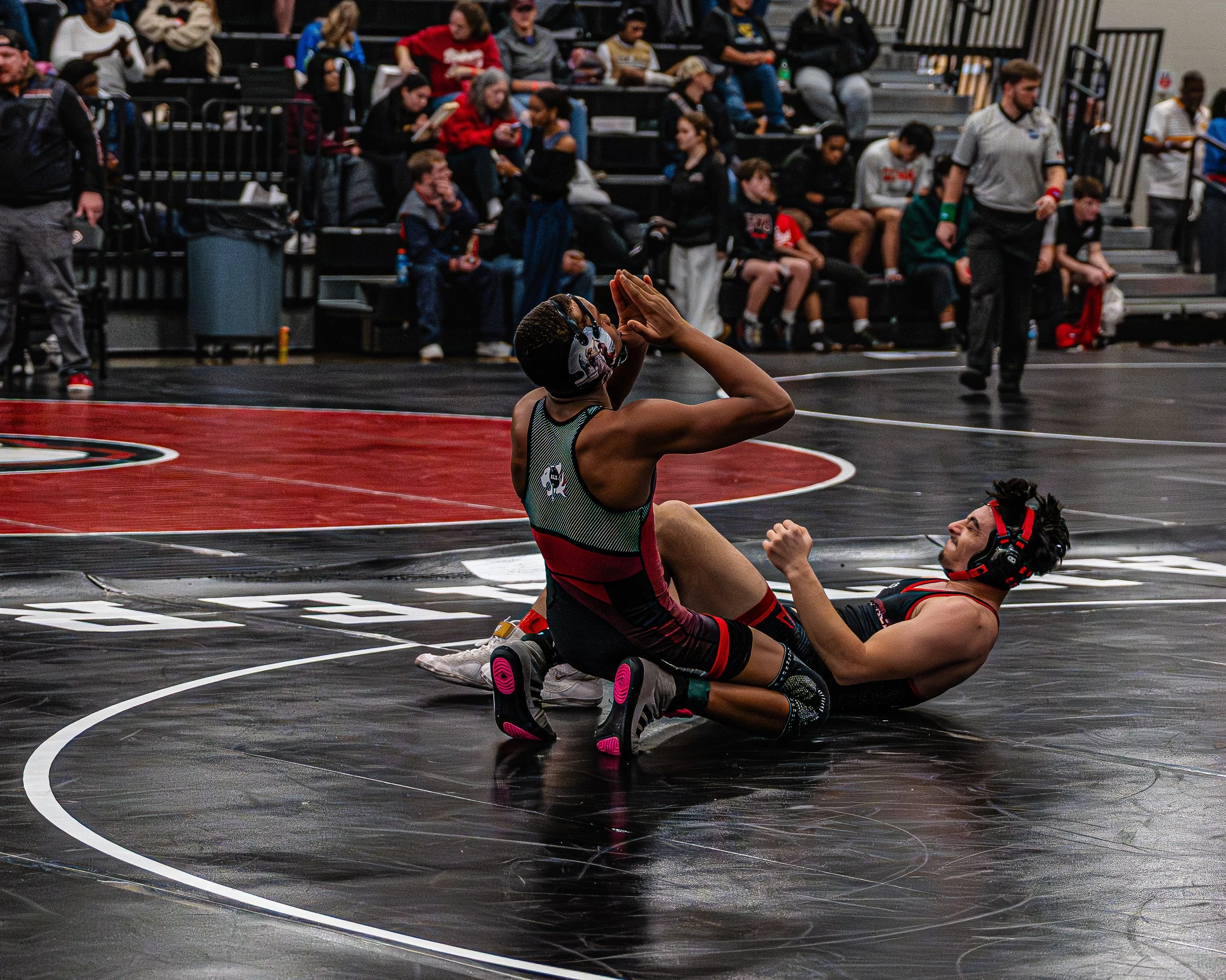 Two wrestlers on a black wrestling mat during a match, with one wrestler on his back and the other on his knees making a gesture, while spectators watch in the background.
