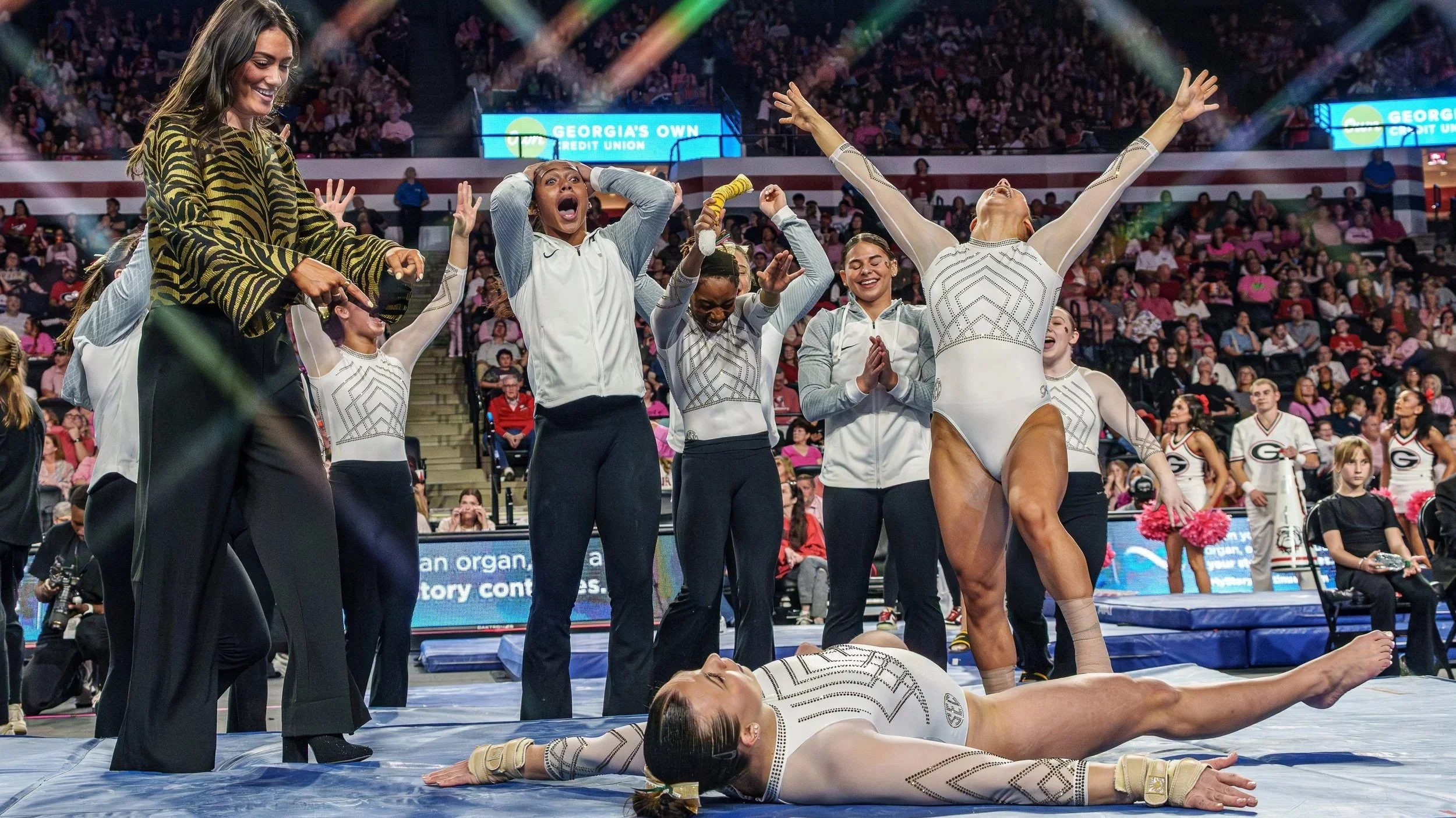 Gymnastics team celebrates on the competition floor, with one gymnast lying on the ground in front and other gymnasts and coaches standing behind her, cheering and clapping, in an arena with audience watching.